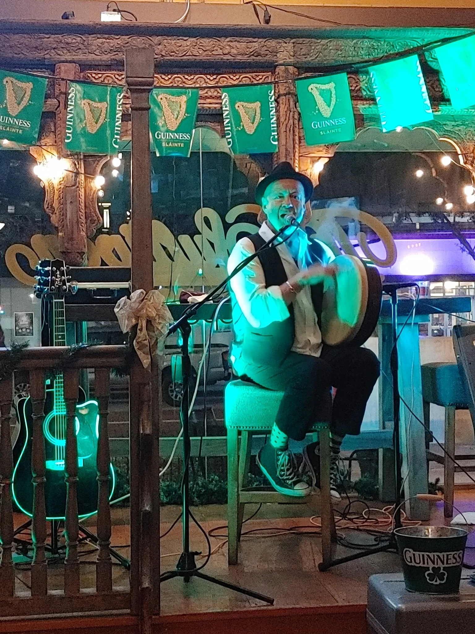 A person dressed in a white shirt with black suspenders, a black hat, and zebra-patterned shoes, playing a drum set at an outdoor event with Guinness banners and glowing neon guitar decorations.