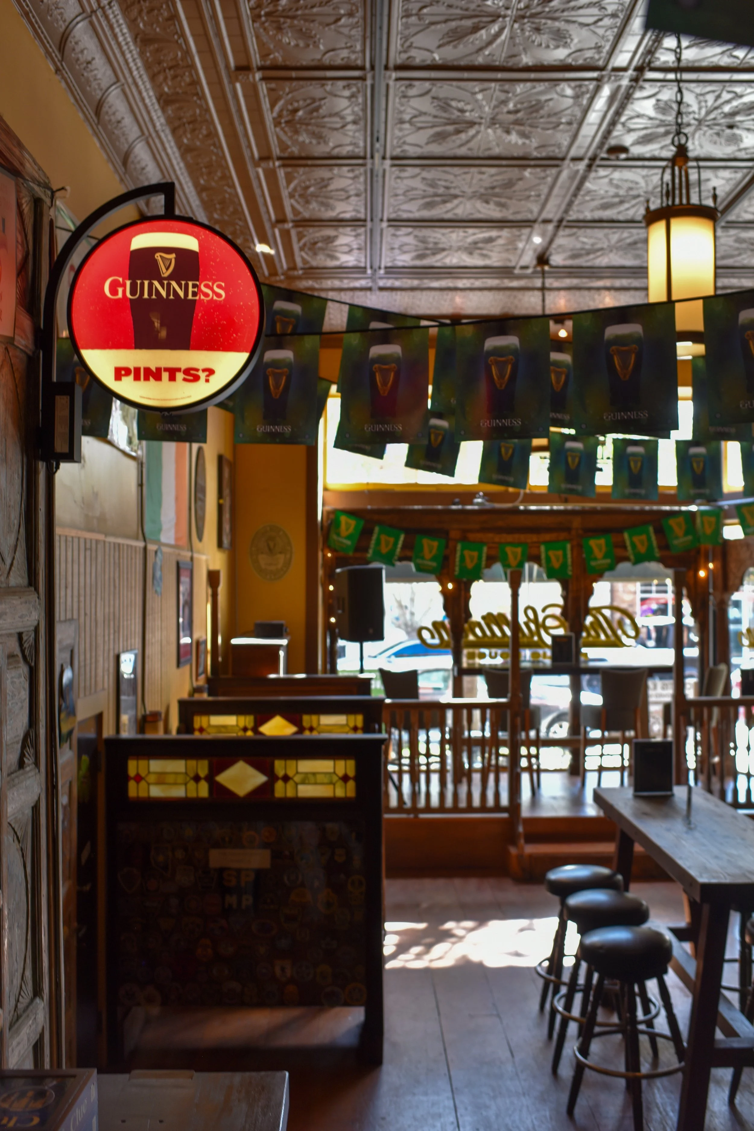 Interior of a pub or bar decorated for St. Patrick's Day, with Guinness-themed banners and flags hanging from the ceiling, and a sign asking 'Pints?' in the Irish Guinness logo style.