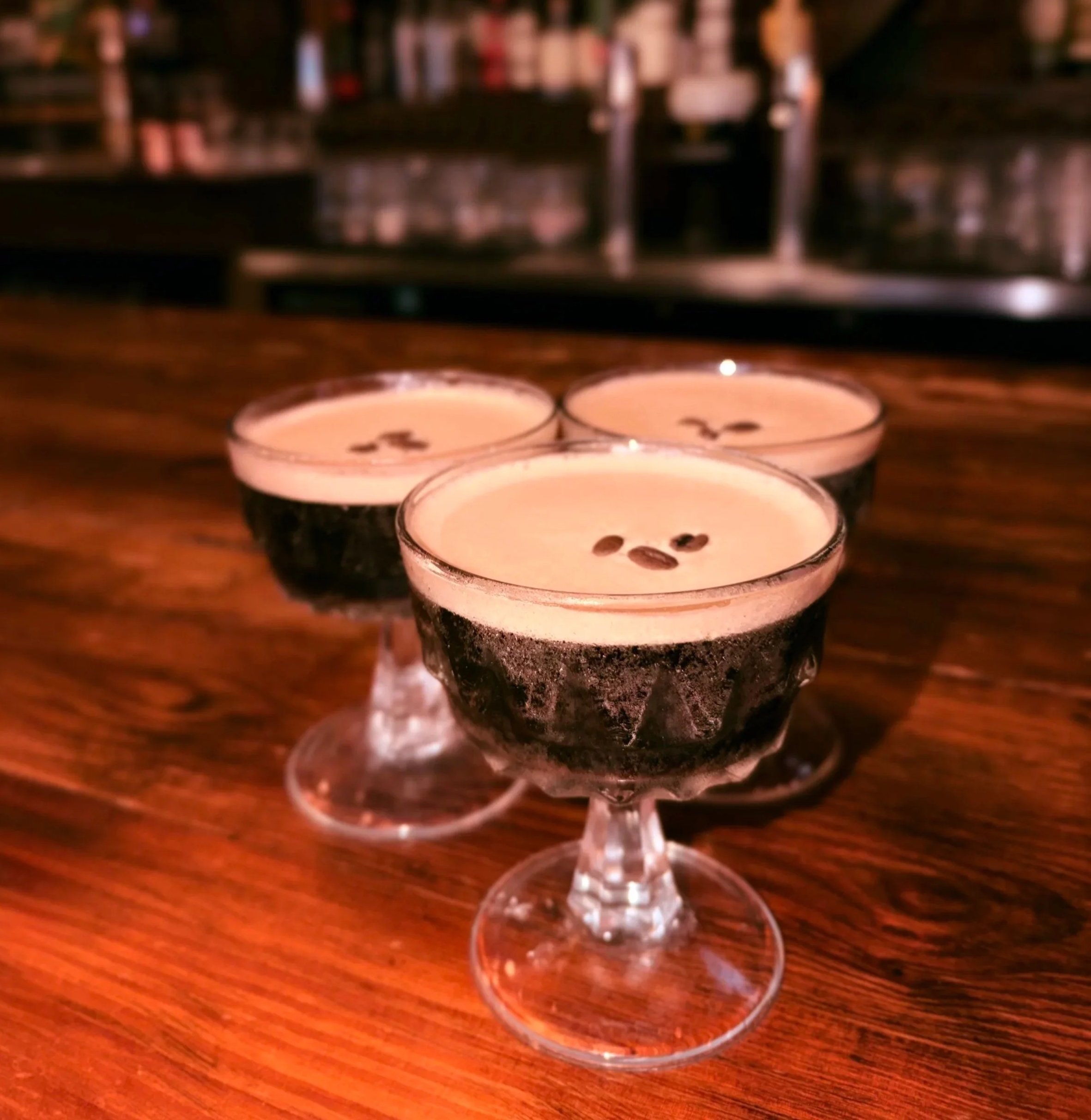Three glasses of coffee with frothy top and coffee beans on the surface, on a wooden bar counter.