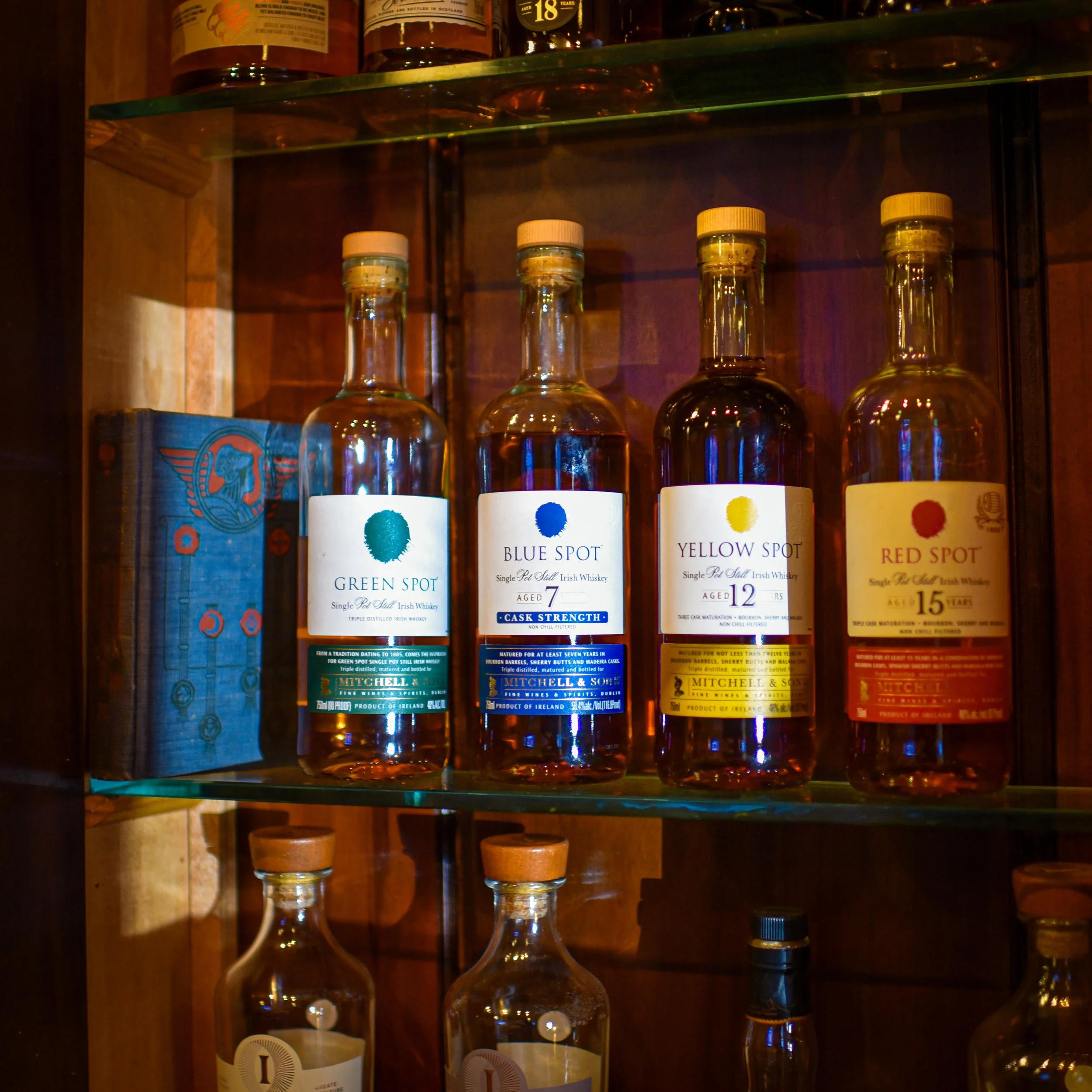 Four bottles of Irish whiskey labeled Green Spot, Blue Spot, Yellow Spot, and Red Spot on a glass shelf in a wooden cabinet.