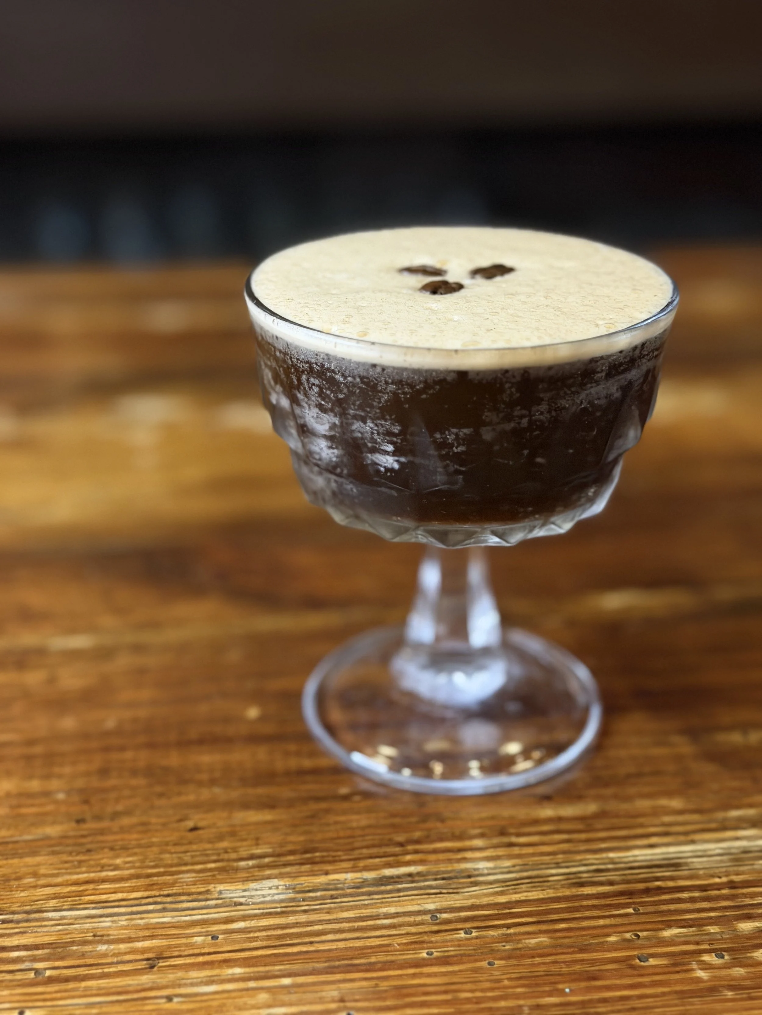 A cocktail drink in a stemmed glass with a frothy top and three coffee beans garnishing the foam, placed on a wooden table.
