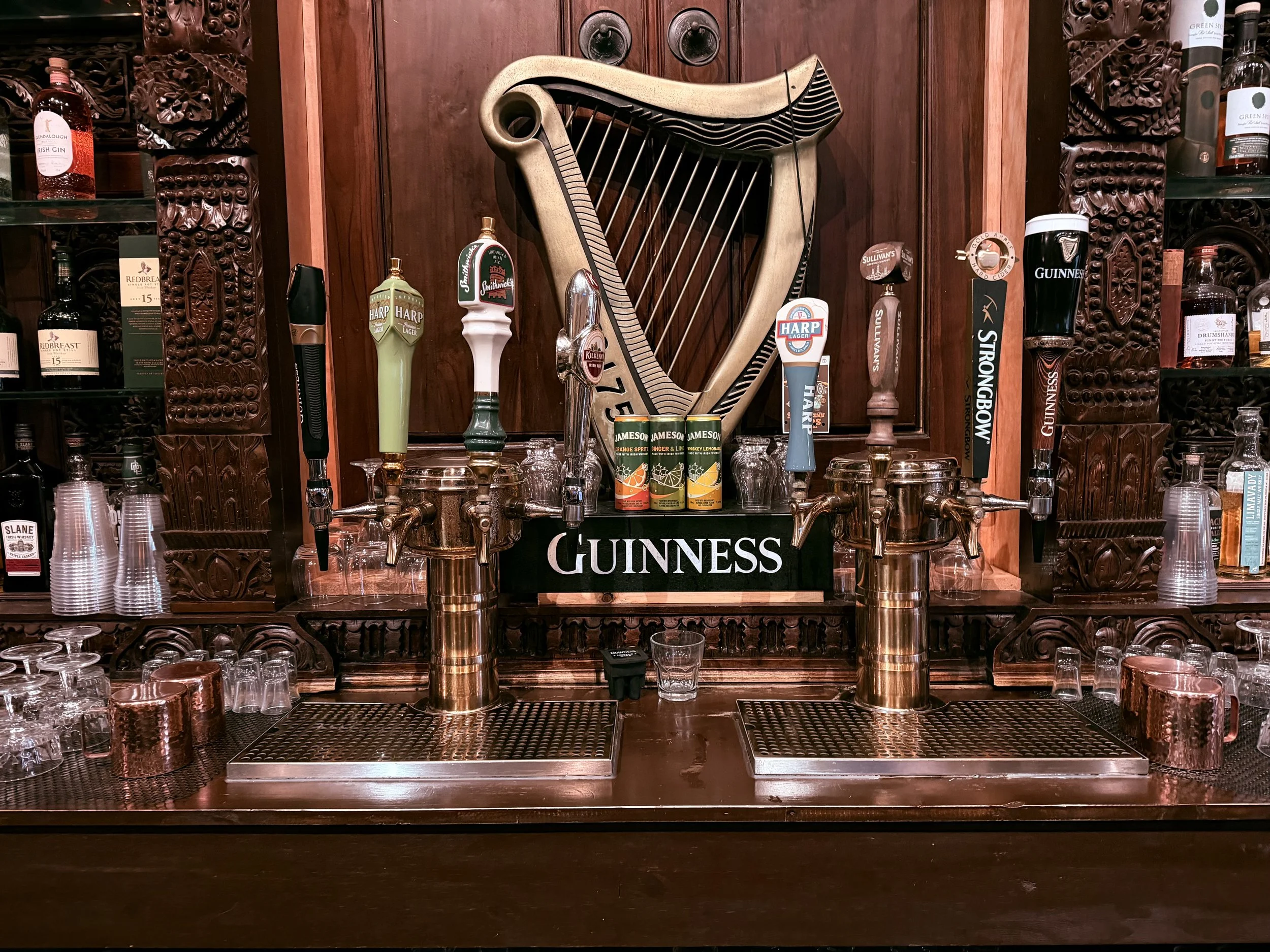 A bar counter with Guinness branding, featuring multiple beer taps, glasses, and liquor bottles, with a harp wall decor behind.