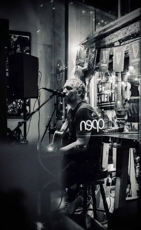 A man with curly hair playing an acoustic guitar and singing into a microphone in a cozy pub or bar with signs and decorations in the background, which include Guinness banners and possibly neon signs.