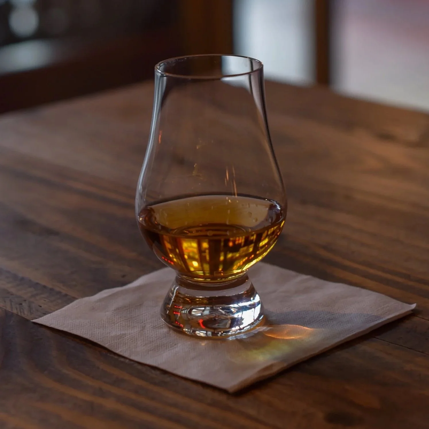 A glass of whiskey on a wooden table with a napkin underneath, reflected light creating patterns on the glass.