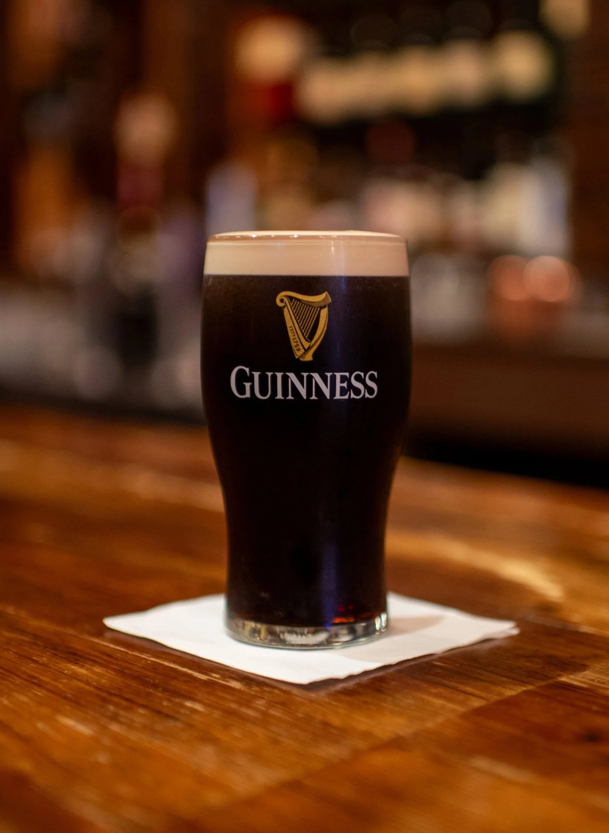 A pint of Guinness stout beer with a creamy head on top, placed on a white napkin on a wooden bar counter.
