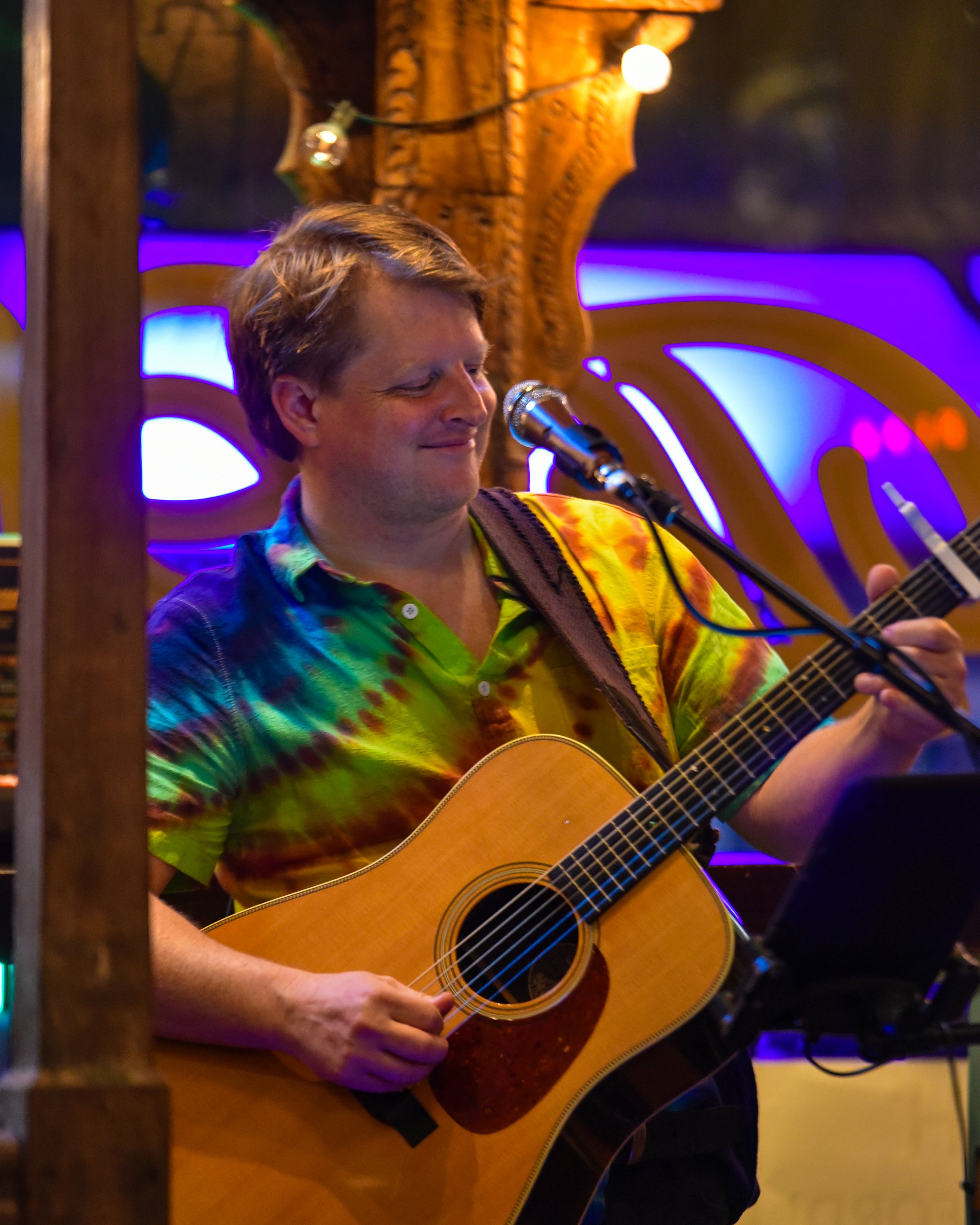 A man playing an acoustic guitar and singing into a microphone in a cozy, colorful venue with blue and purple lighting, wooden decorations, and string lights.