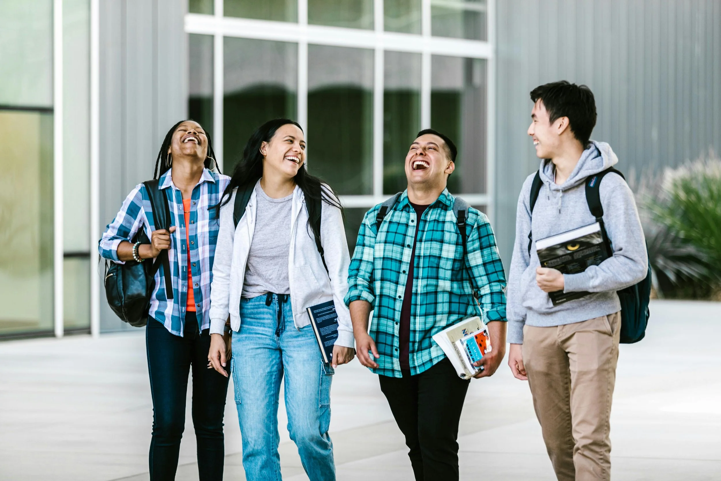 Four diverse college students walking outdoors and laughing together, carrying books and backpacks, in front of a modern building with large windows.
