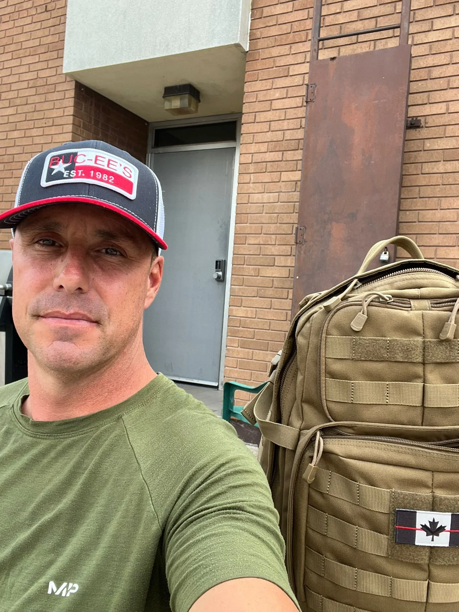 A man taking a selfie outdoors, wearing a green t-shirt and a gray and red BUCEES baseball cap. Behind him are a brick building with a gray door and a lock, a small overhang, and a wall-mounted light. To his right is a tan tactical backpack with a Canadian flag patch on it.