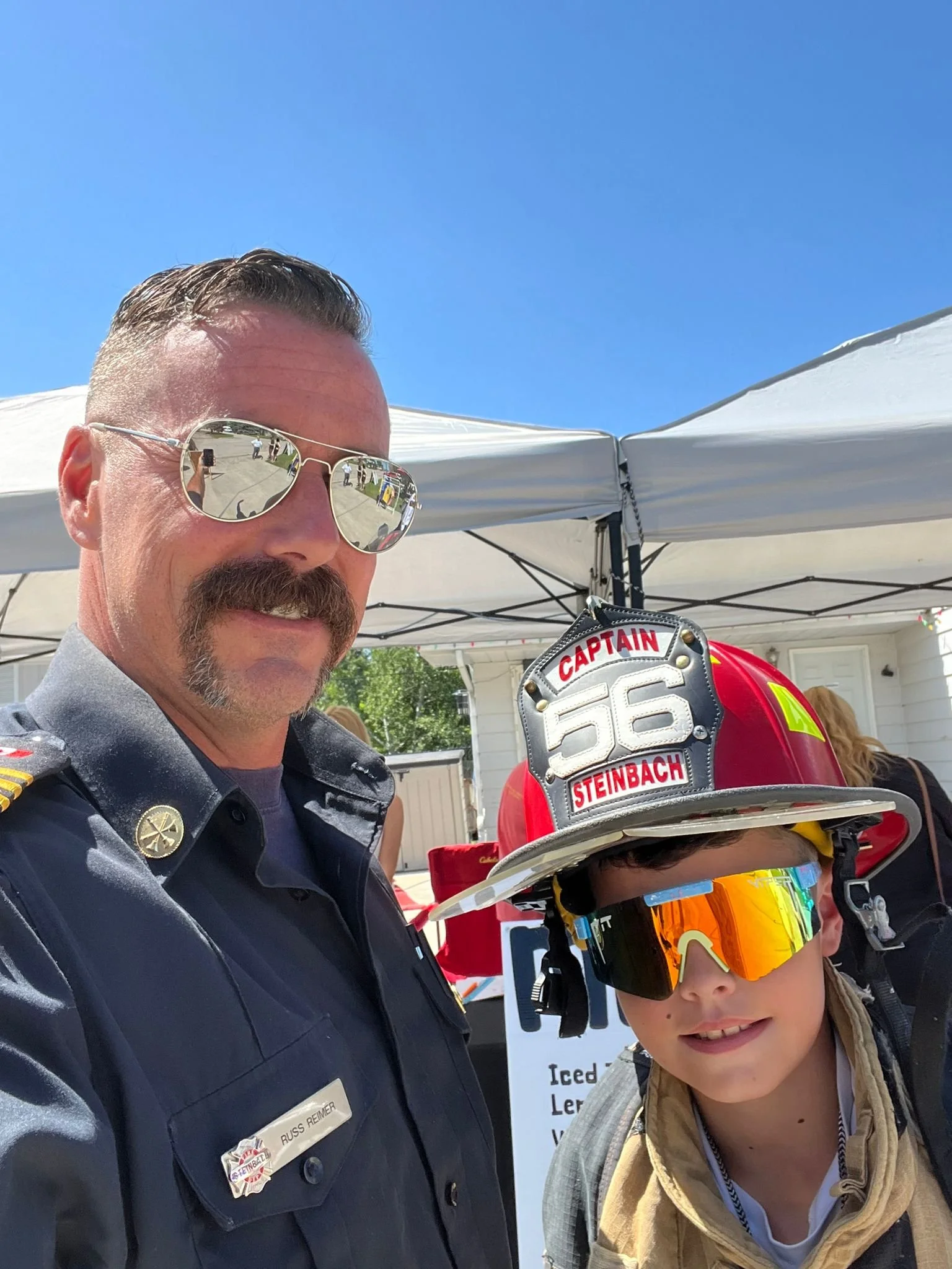 A man dressed as a police officer with sunglasses and mustache, standing next to a young firefighter girl wearing a firefighter's helmet with the words 'Captain Steinbach.' They are outdoors under white tents with a clear blue sky in the background.