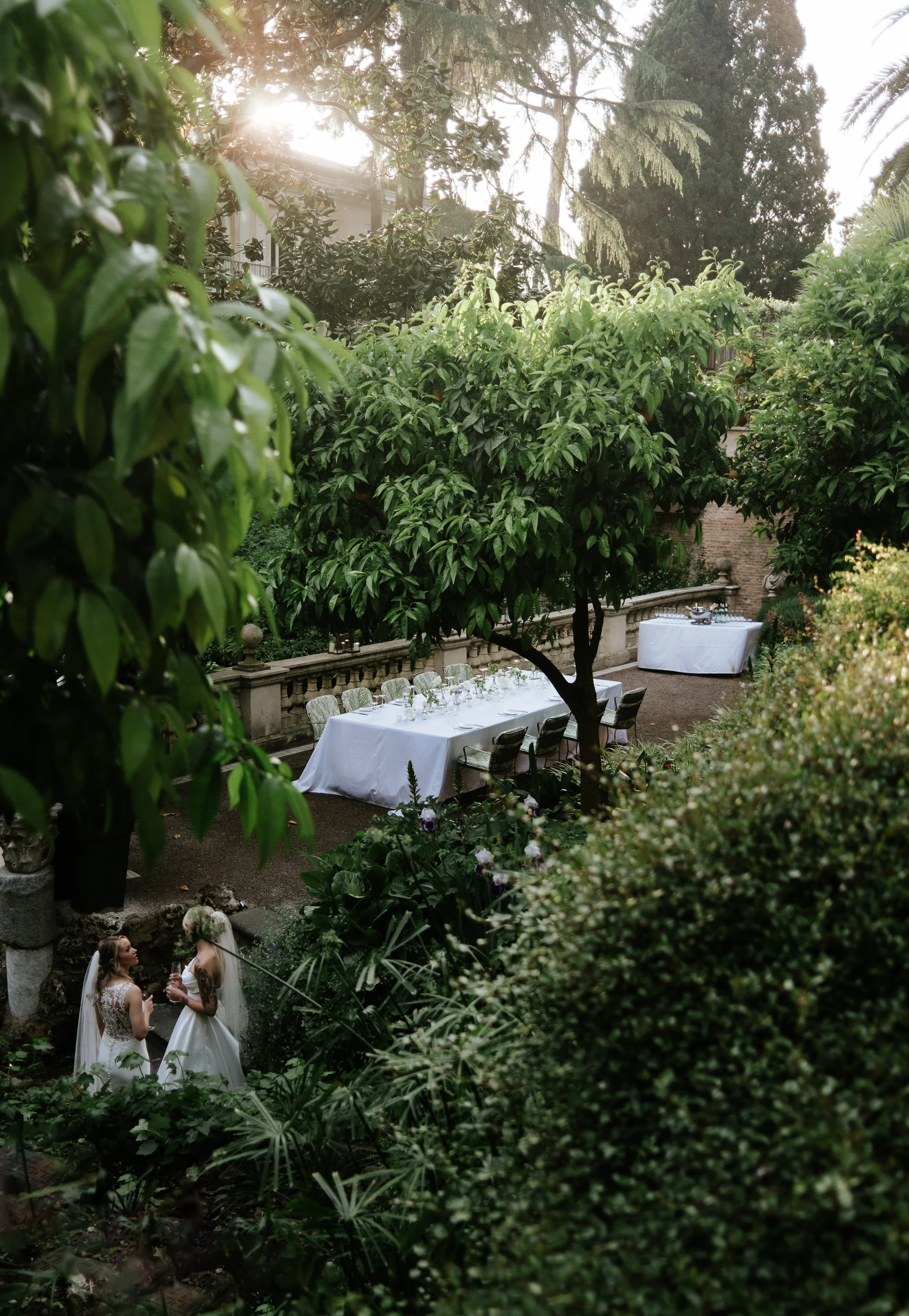 Un giardino verde con una grande tavola imbandita con tovaglia bianca, circondata da sedie, e due donne in abiti da sposa che parlano tra loro.