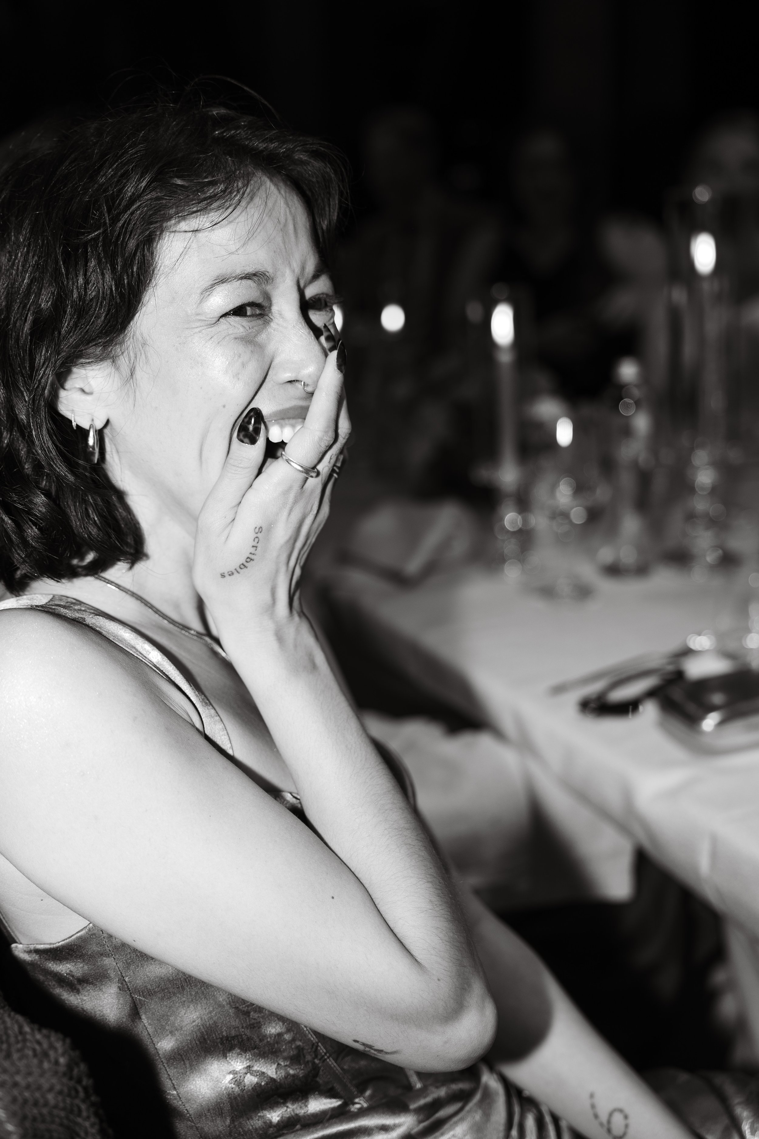 Una donna con capelli ricci scuri sorride mentre copre bocca con mano in una cena, sfondo con tavolo e bicchieri.