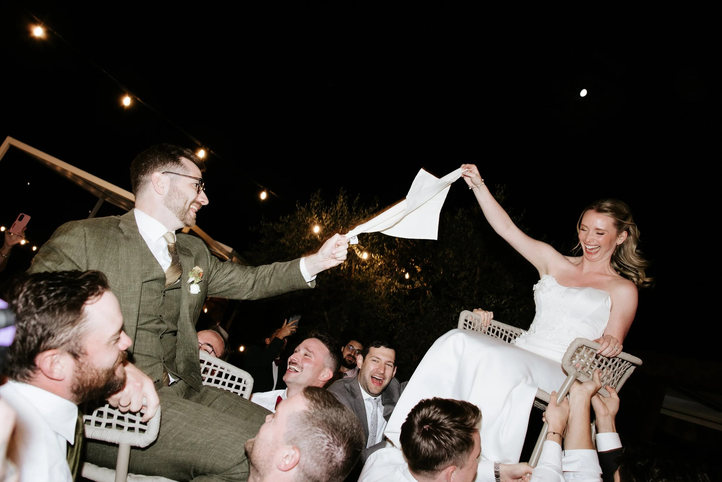 Una coppia di sposi celebra il matrimonio durante una festa all'aperto di notte, con la sposa seduta su una sedia sollevata da amici, mentre il marito le dà il lembo di un tovagliolo, entrambi sorridenti e felici.