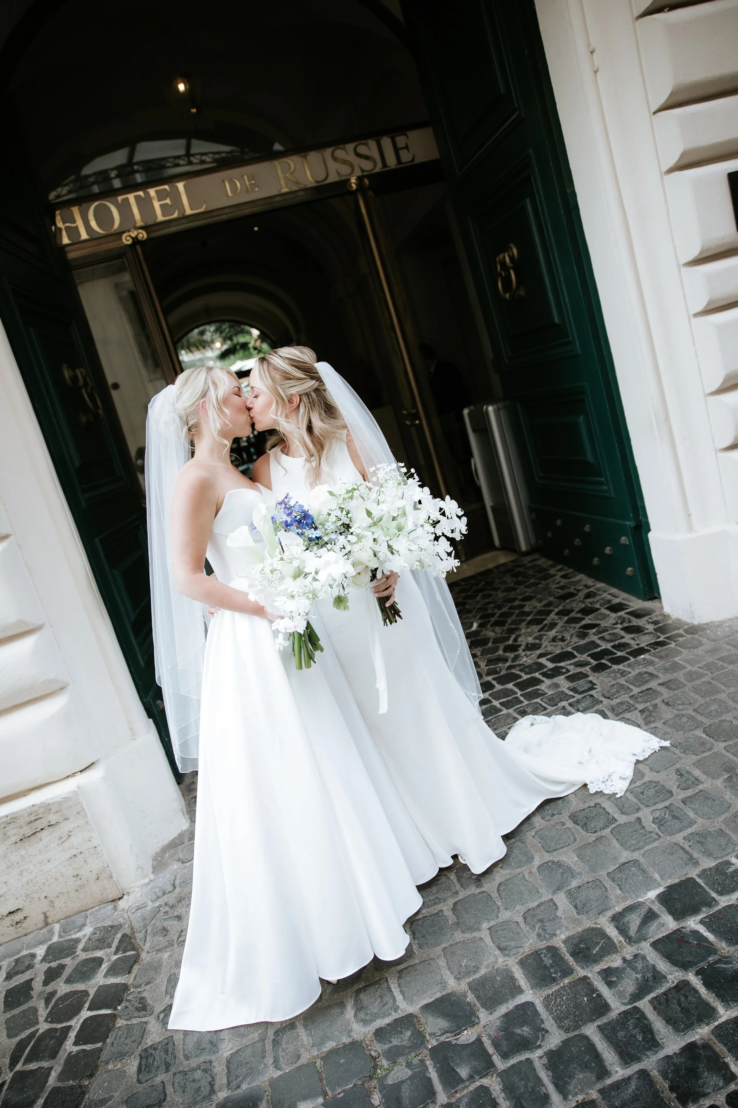 Due spose in abiti da matrimonio si baciano davanti all'ingresso dell'Hotel de Russie a Roma, con bouquet di fiori bianchi e decorazioni eleganti.