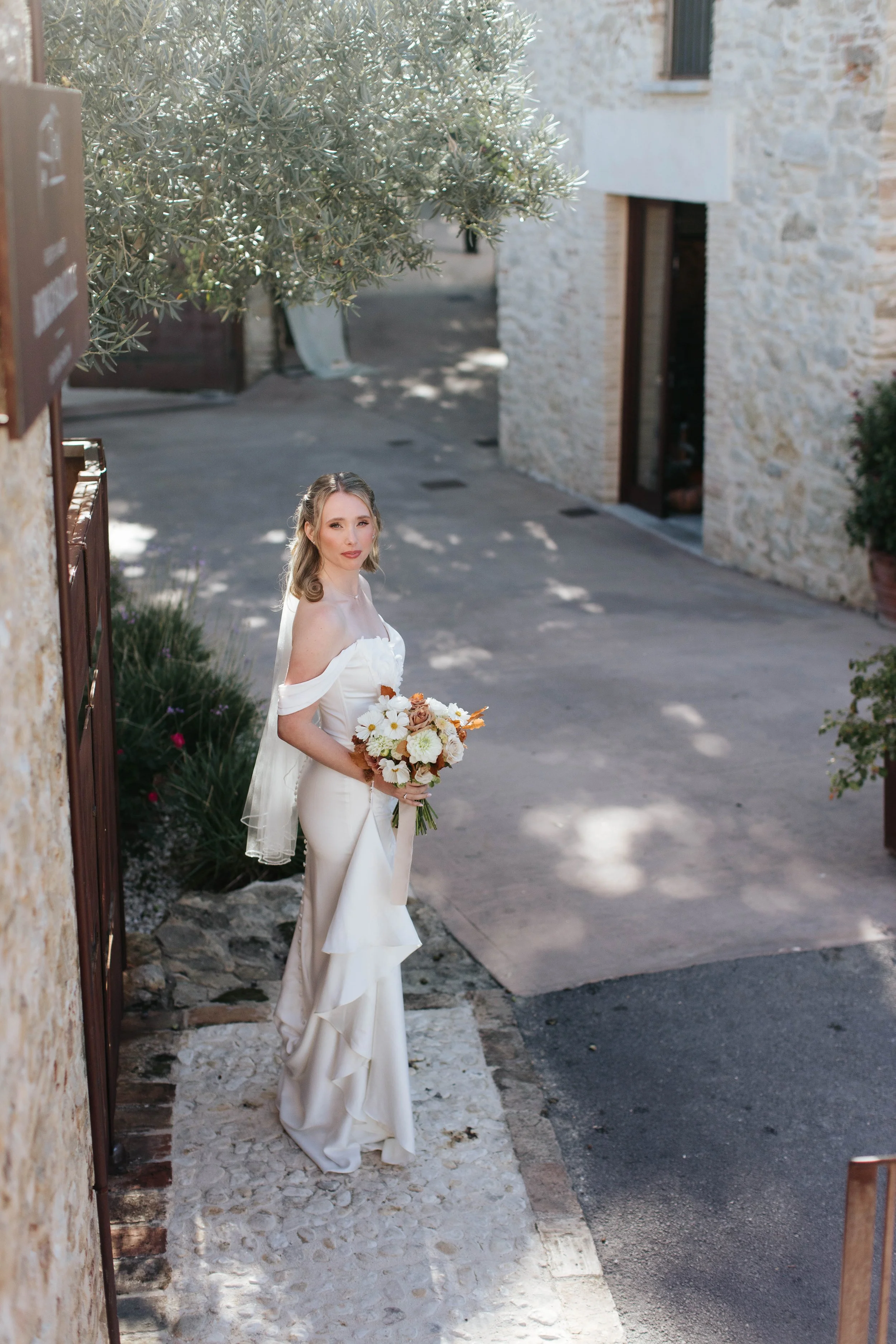 Una donna con abito da sposa bianco, che tiene un mazzo di fiori, si trova in un vicolo con case di pietra e piante, illuminata dalla luce del sole
