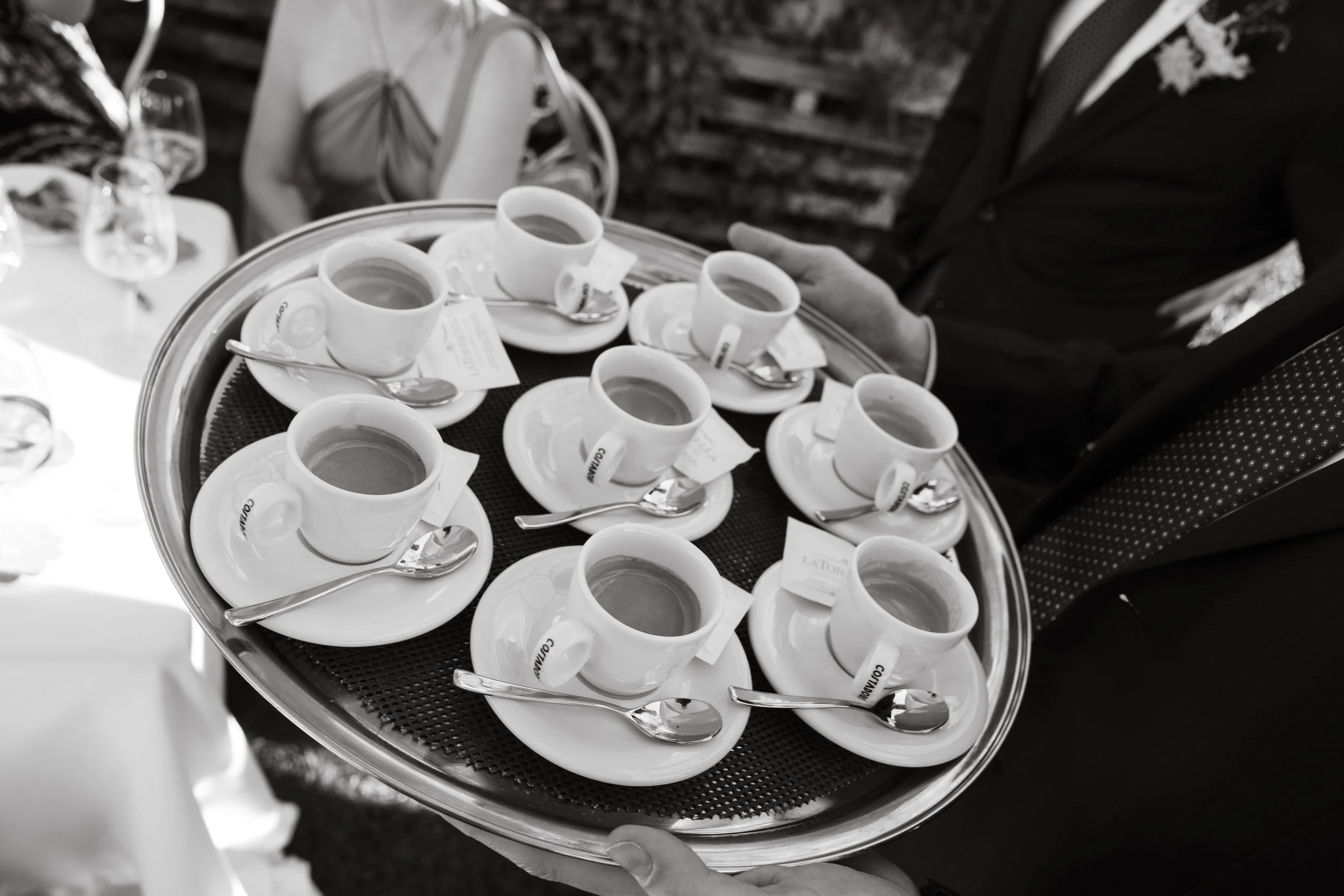 Un portafrutta con diverse tazze di caffè e cucchiaini, sorretto da una persona in abito elegante, presumo a una cerimonia o evento formale.
