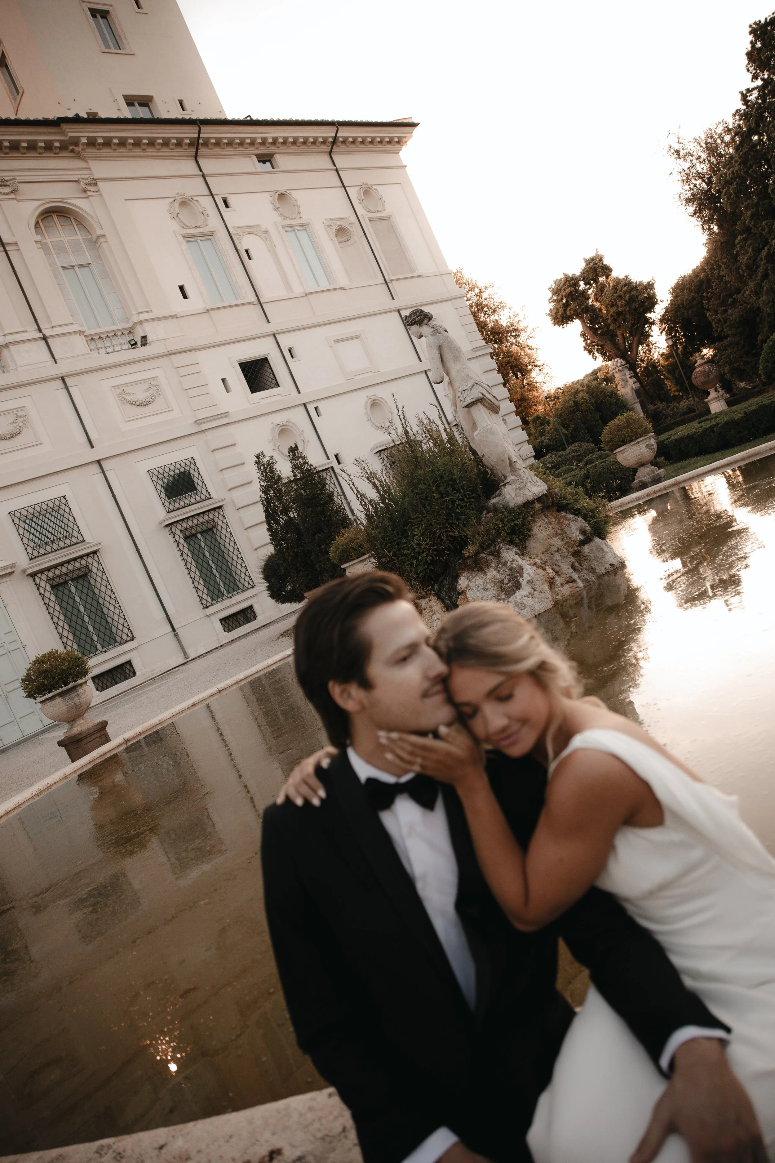 Una coppia di sposi in un abbraccio romantico davanti a uno stagno con statua e giardino in un edificio storico al tramonto.