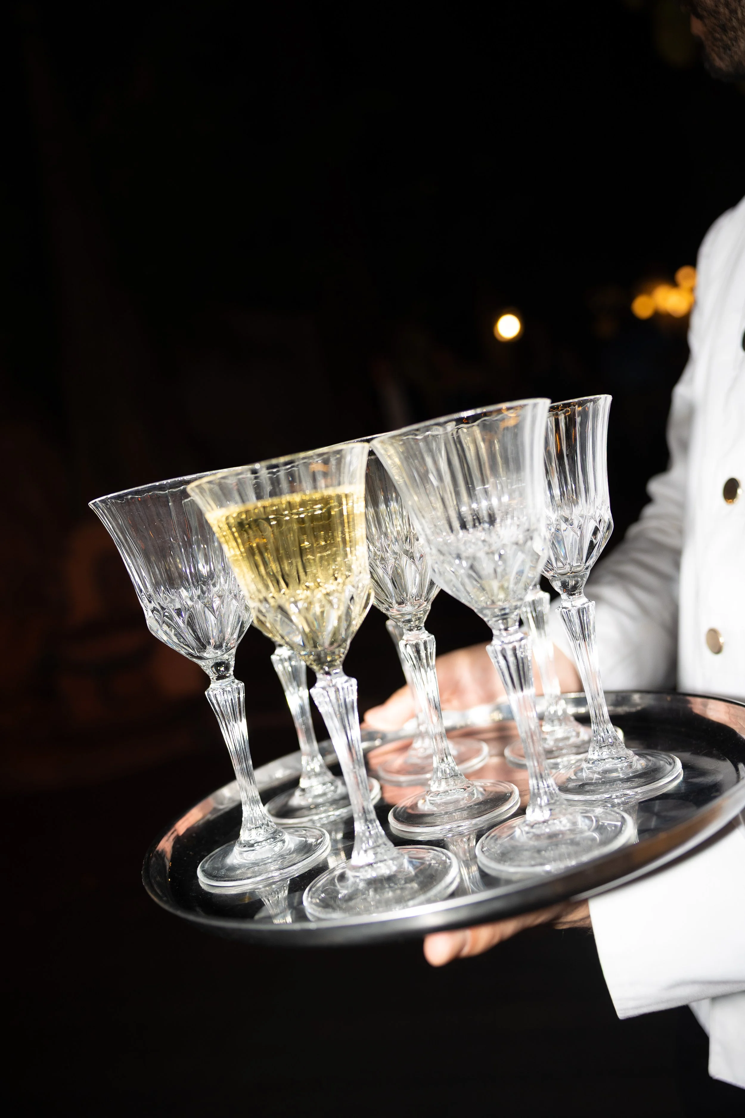 Un cameriere che tiene un vassoio con sei bicchieri di cristallo, uno di questi riempito di champagne, in un evento serale.