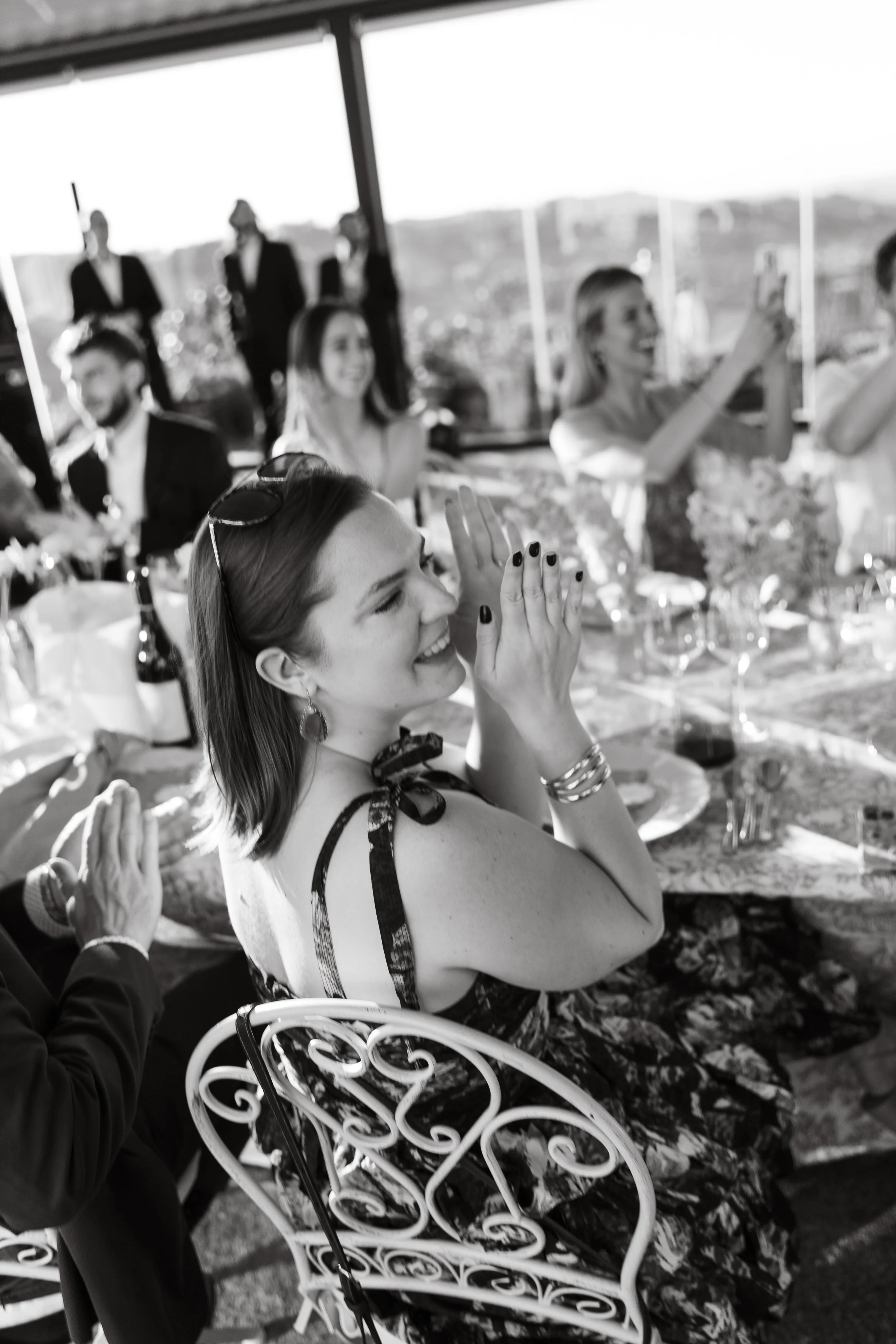Persone sedute a un banchetto durante un evento, con alcune che scattano fotografie e altre che ridono, in un ambiente con grandi finestre e vista panoramica.