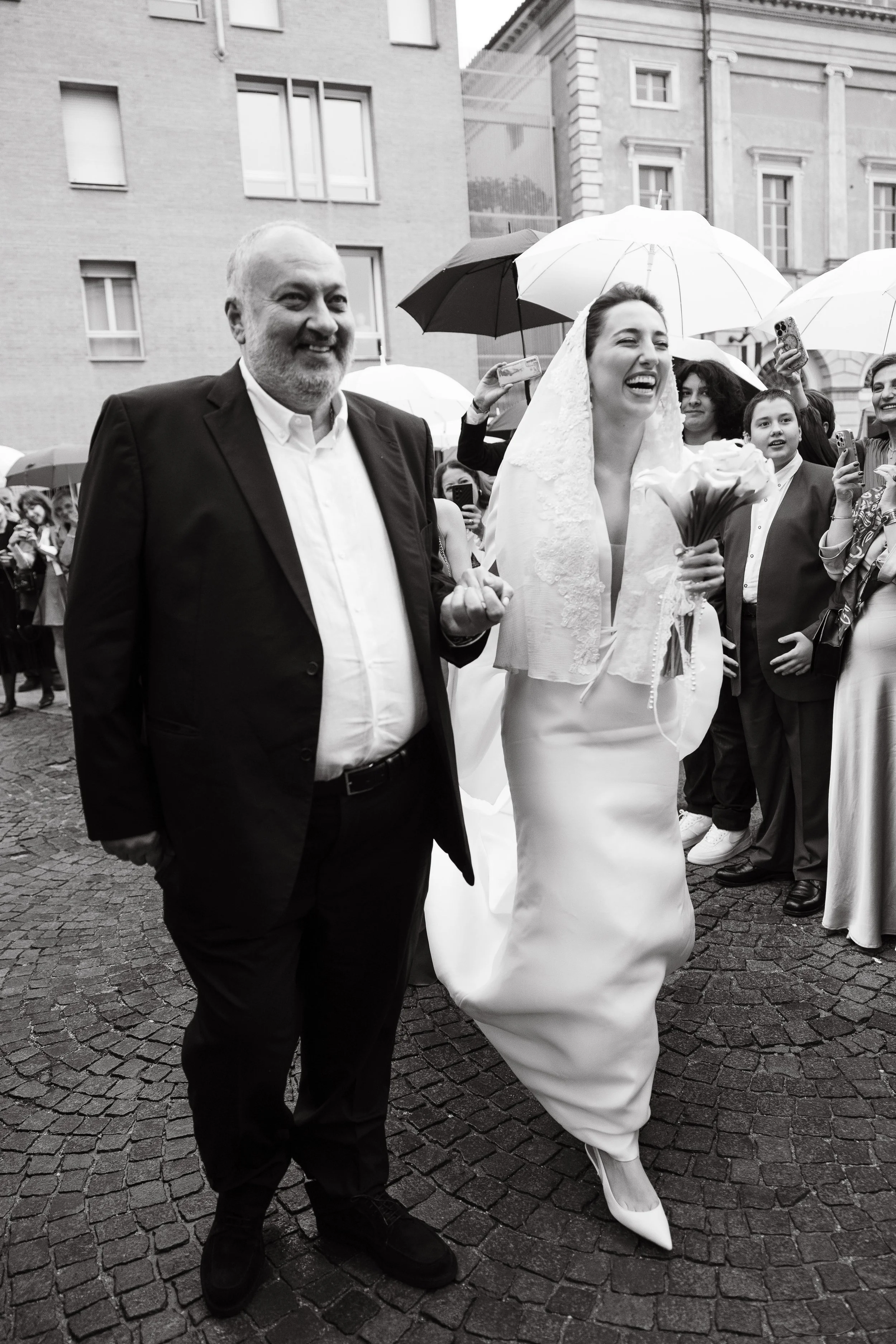 Bride and her dad walking towards the church before the ceremony. She smiles and they are happy with the guests. Duomo di Alba, Piedmont.