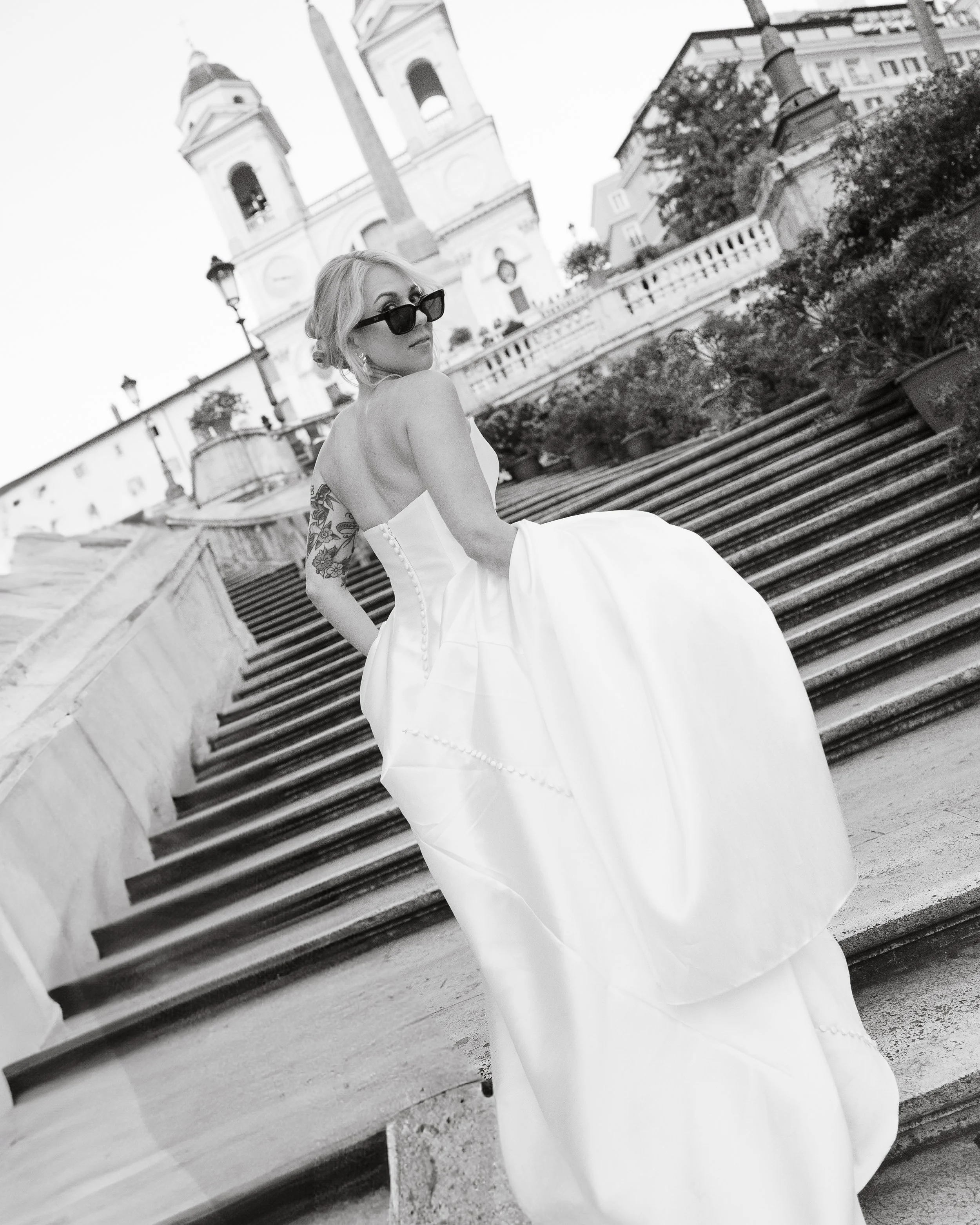 Una donna vestita con un abito bianco lungo e con occhiali da sole neri cammina sulle scale di una chiesa in una zona urbana, in bianco e nero.