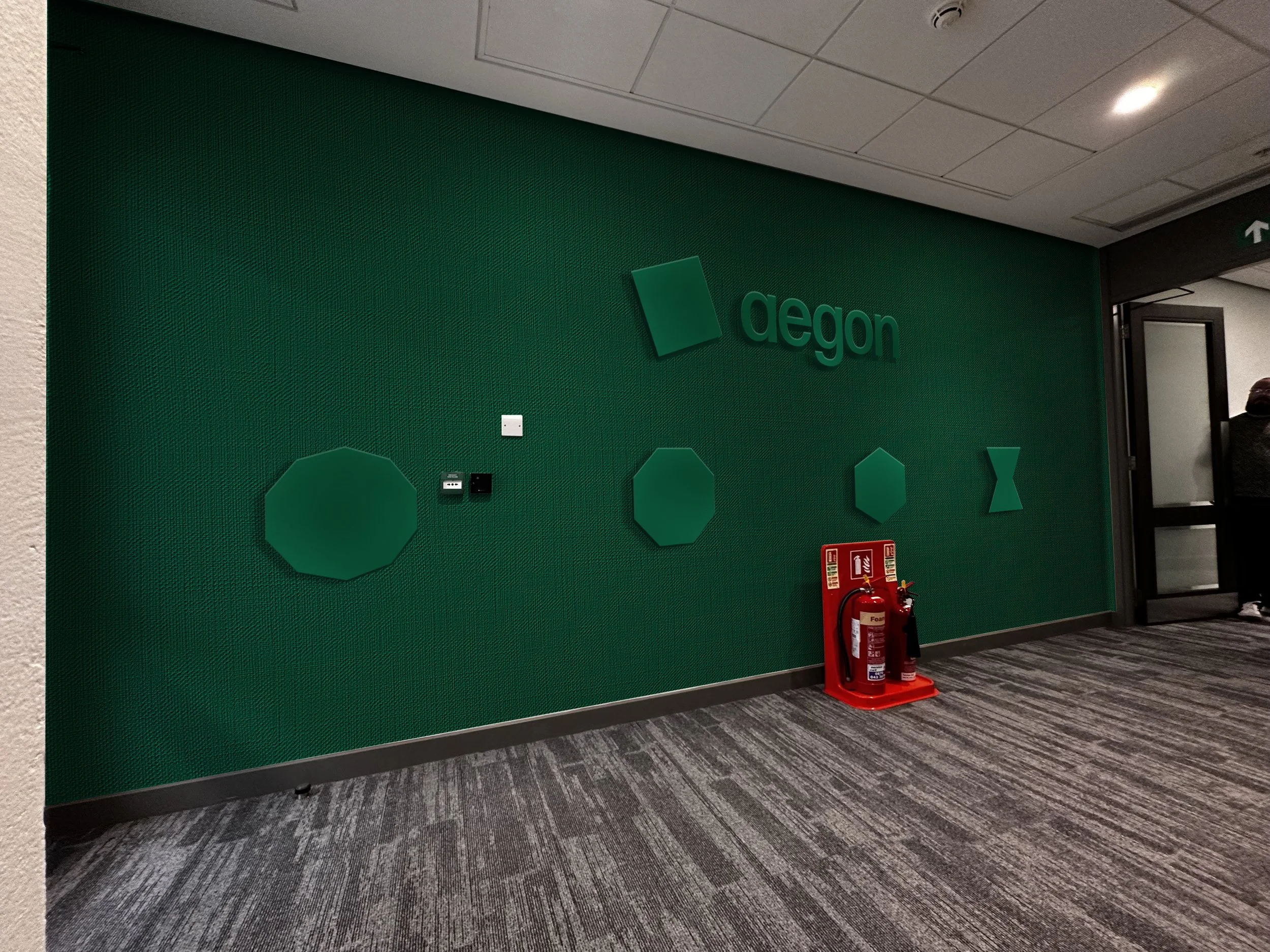 Green wall with the word 'Aegon' and its logo in a building interior, fire extinguisher and fire alarm on the floor, and a person near an open door to the right.
