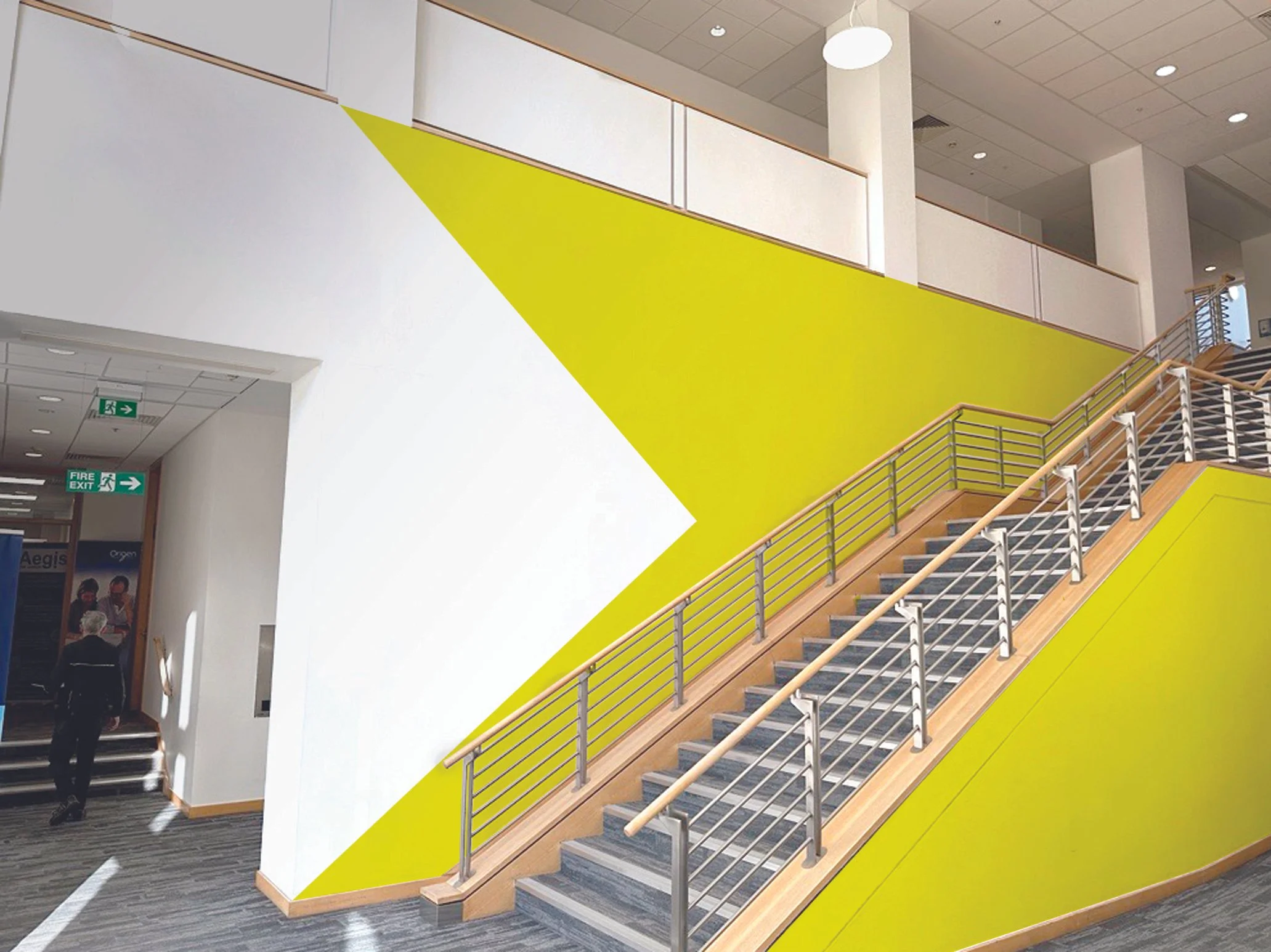 Interior of a modern building with a staircase, a yellow and white wall design, and a person walking near an exit sign.