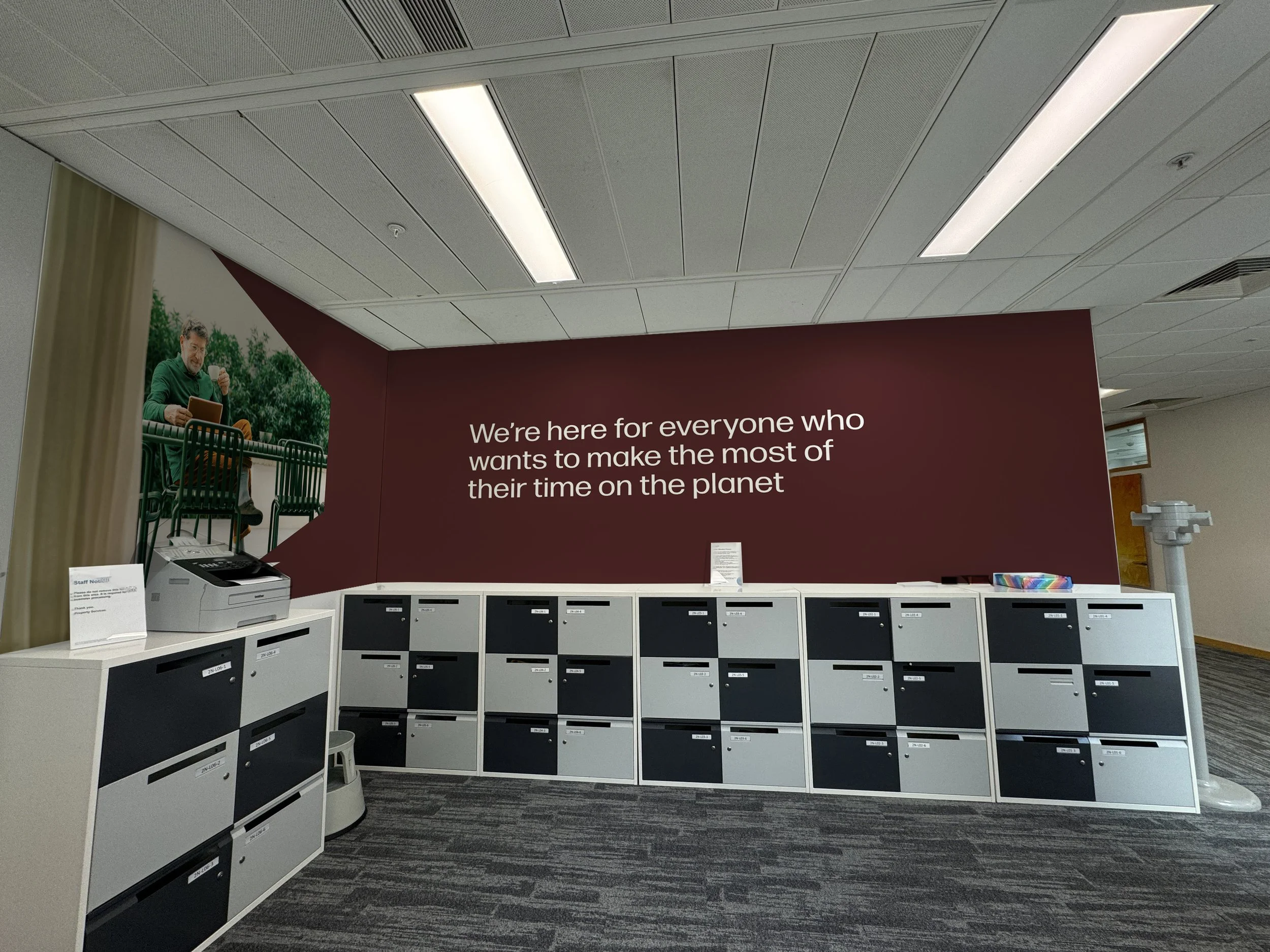 An indoor scene showing a row of white and black mail or storage lockers on a carpeted floor, with a maroon wall behind them displaying white text: 'We're here for everyone who wants to make the most of their time on the planet.' To the left, there i
