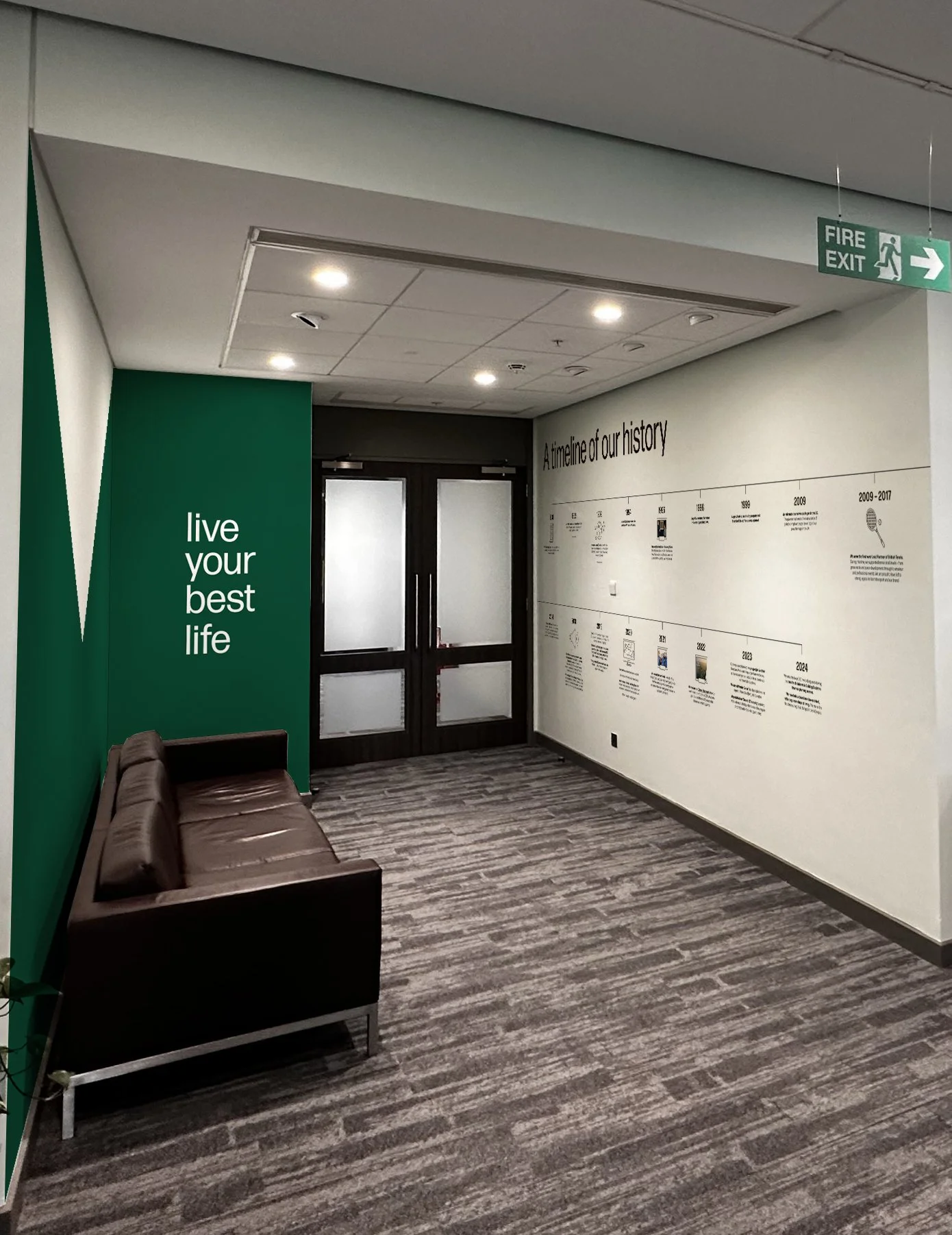 Interior hallway with a green accent wall that reads 'live your best life', a brown leather bench, double glass doors, a timeline of history on the wall, and a fire exit sign hanging from the ceiling.