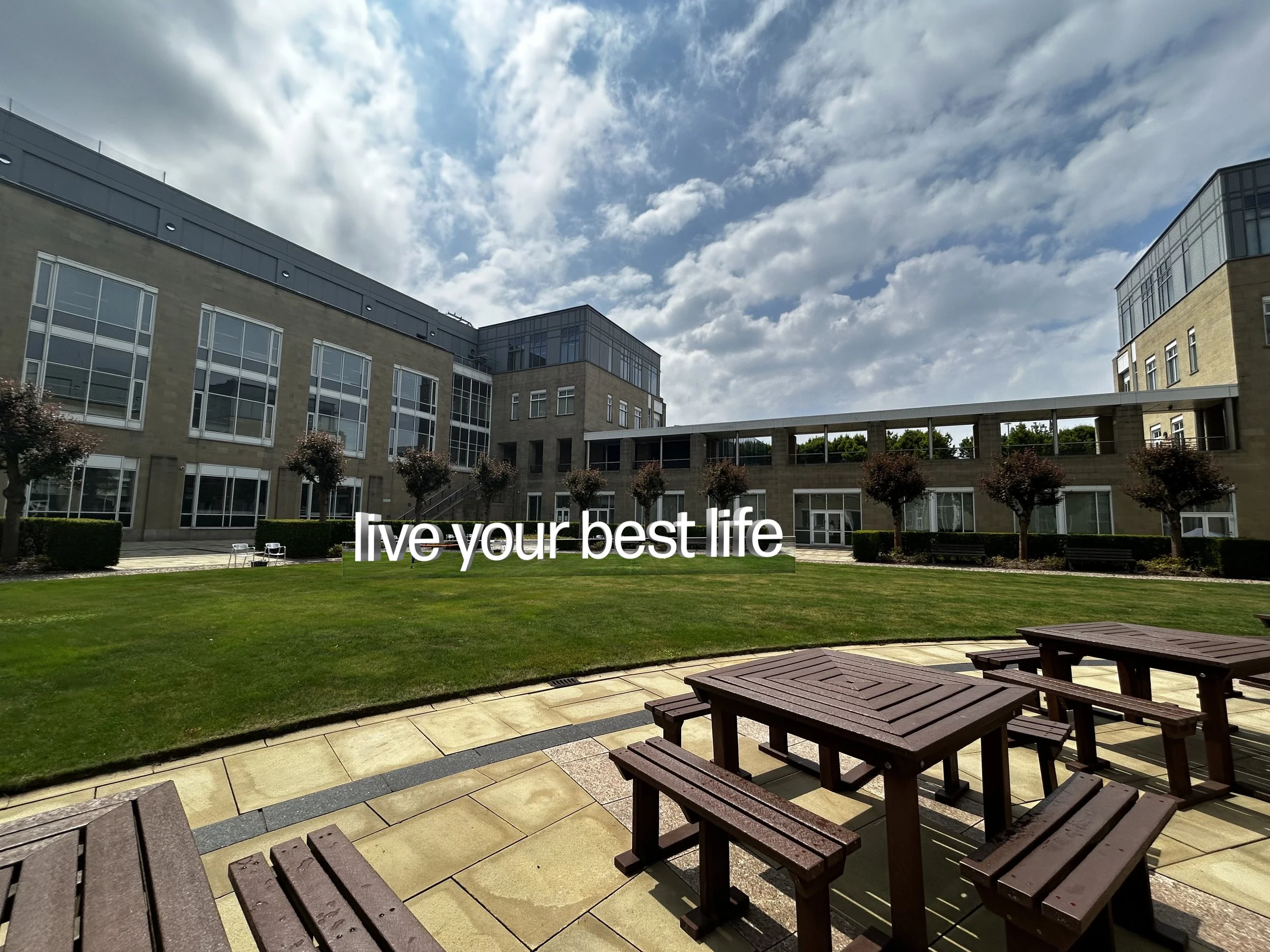 An outdoor courtyard with wooden picnic tables, a green lawn with small trees, and a modern building with large windows under a partly cloudy sky. Text overlay says 'live your best life.'