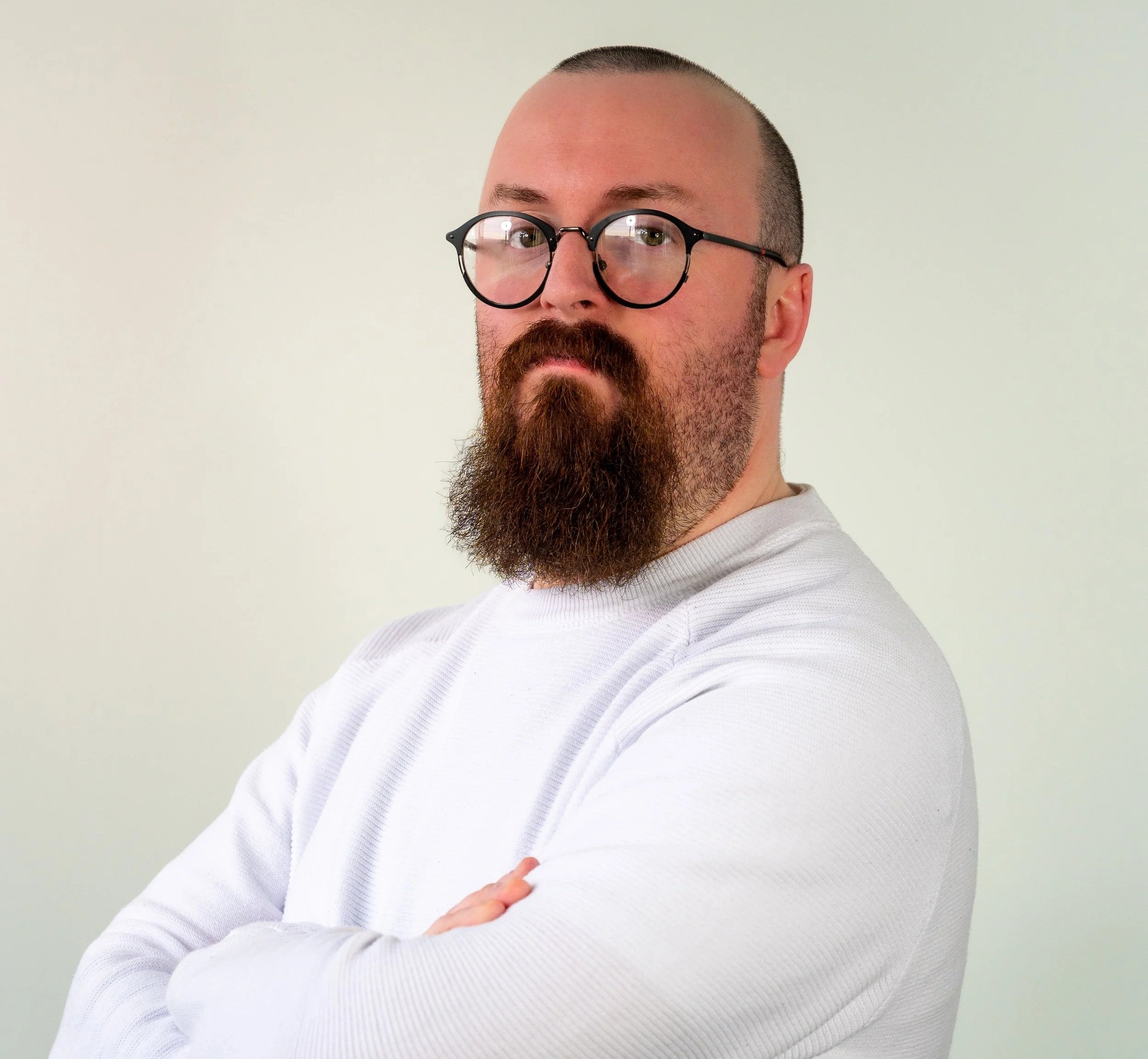 A man with a beard, glasses, and crossed arms wearing a white long sleeve shirt, posing against a plain background.