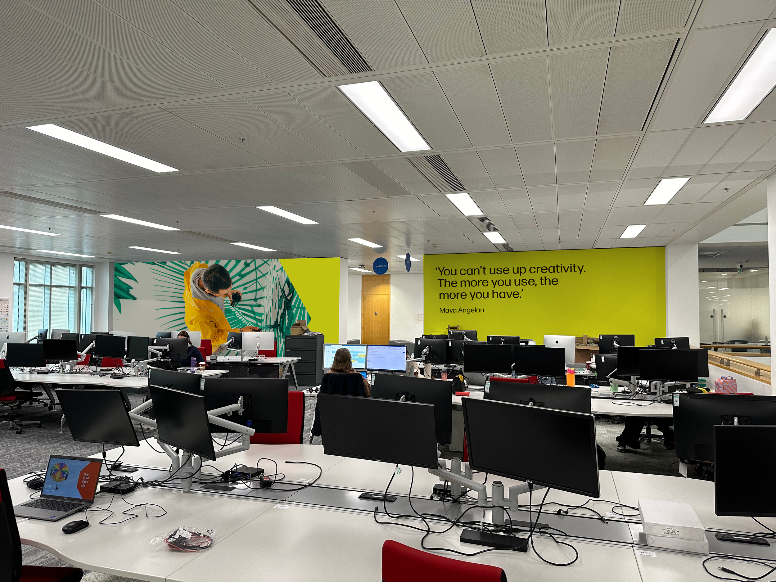 Modern office space with multiple desks equipped with computer monitors, some workstations have laptops, and a few employees working at their desks. The back wall features a large mural of a person in a yellow jacket with green tropical leaves, and a