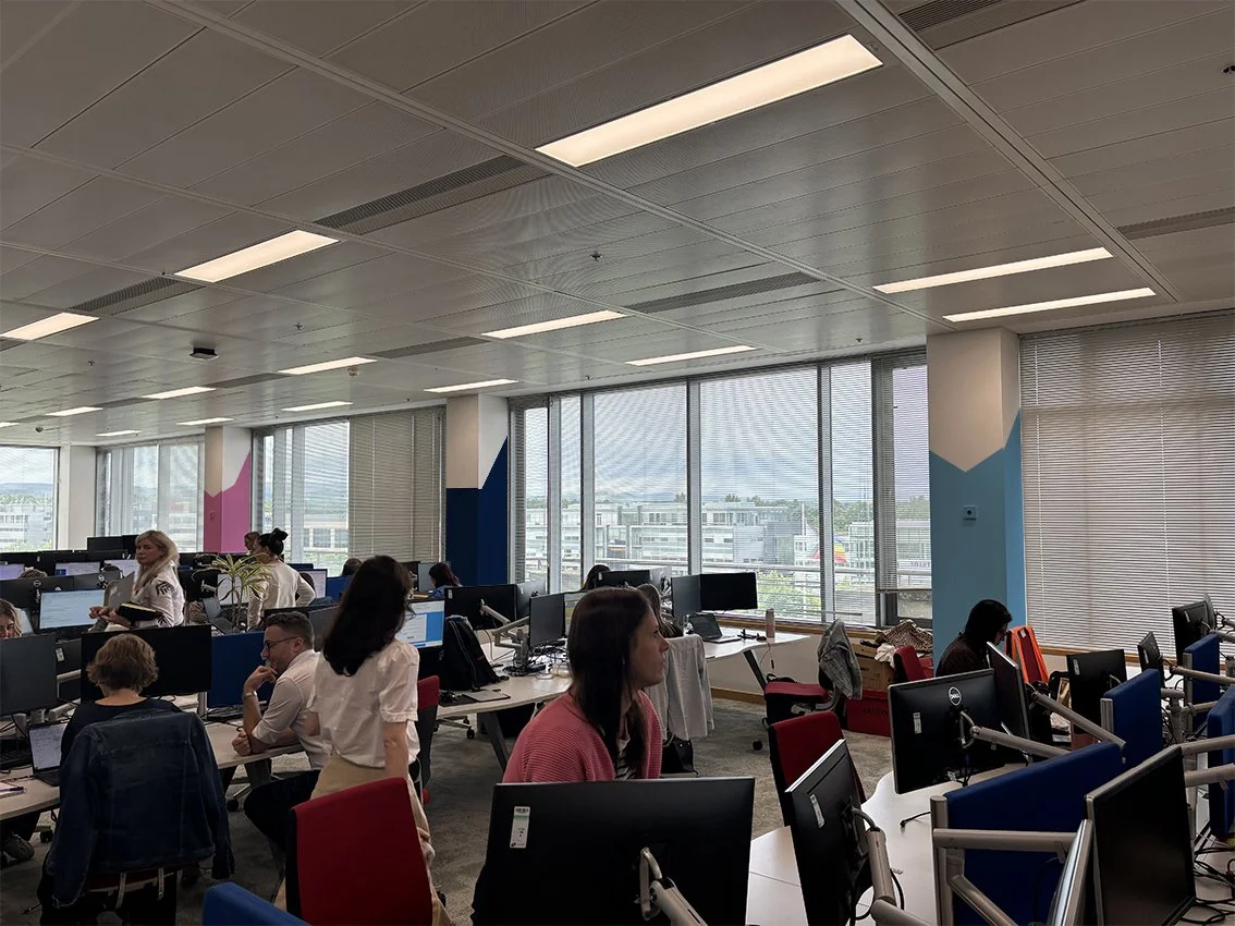 A busy office with multiple workers at desks, some standing and some sitting, with large windows in the background showing a cityscape. The office has color accents on the walls and is well-lit with ceiling lights.