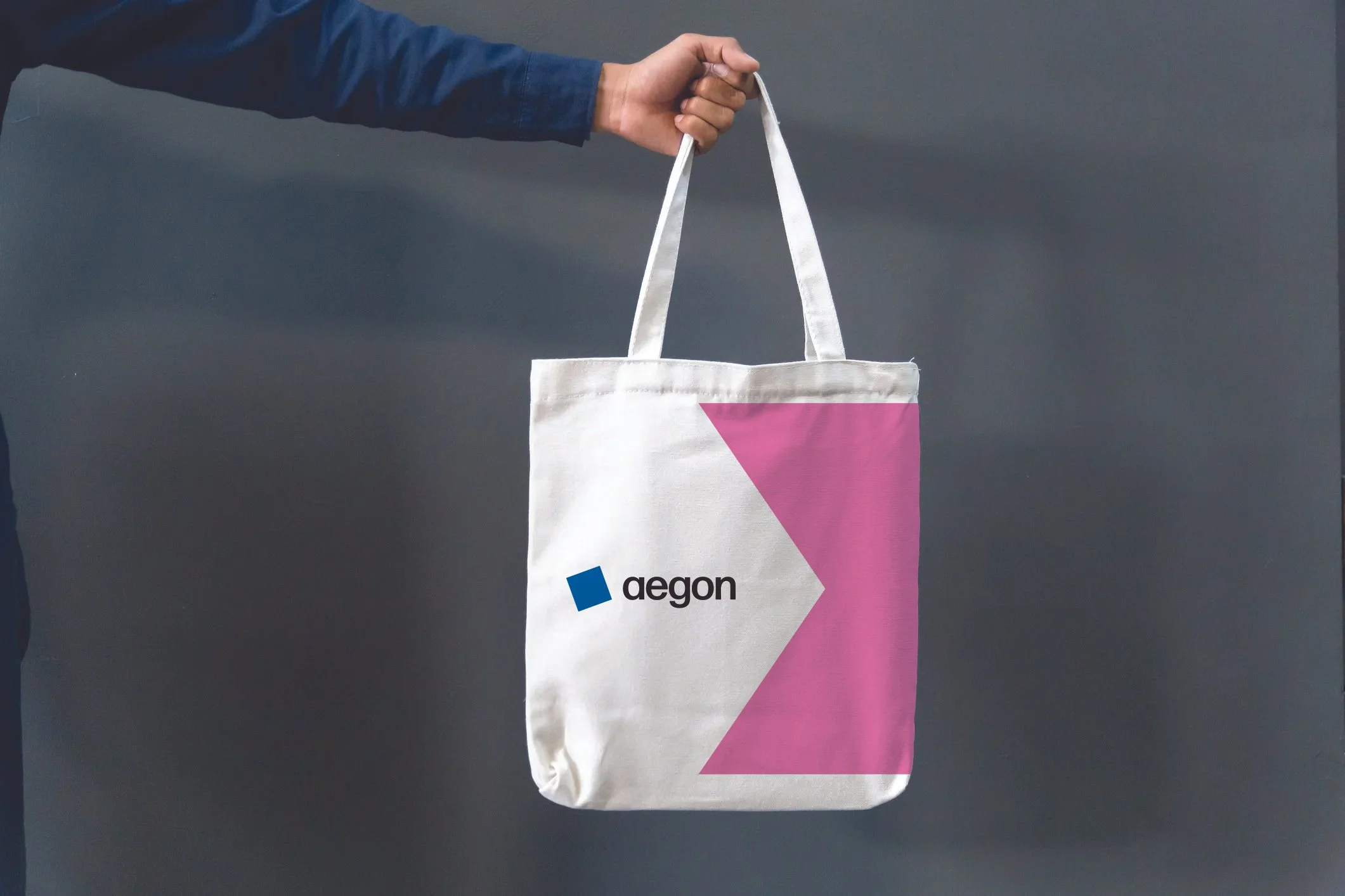 A person holding a white tote bag with a pink geometric design and a logo that says 'aegon' on it, against a dark grey background.