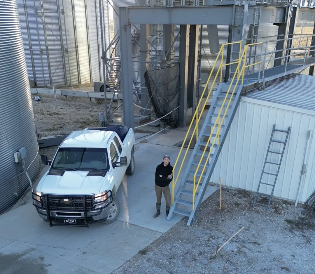 A person standing next to a white pickup truck on a concrete pad near industrial silos and metal stairs at an industrial site.