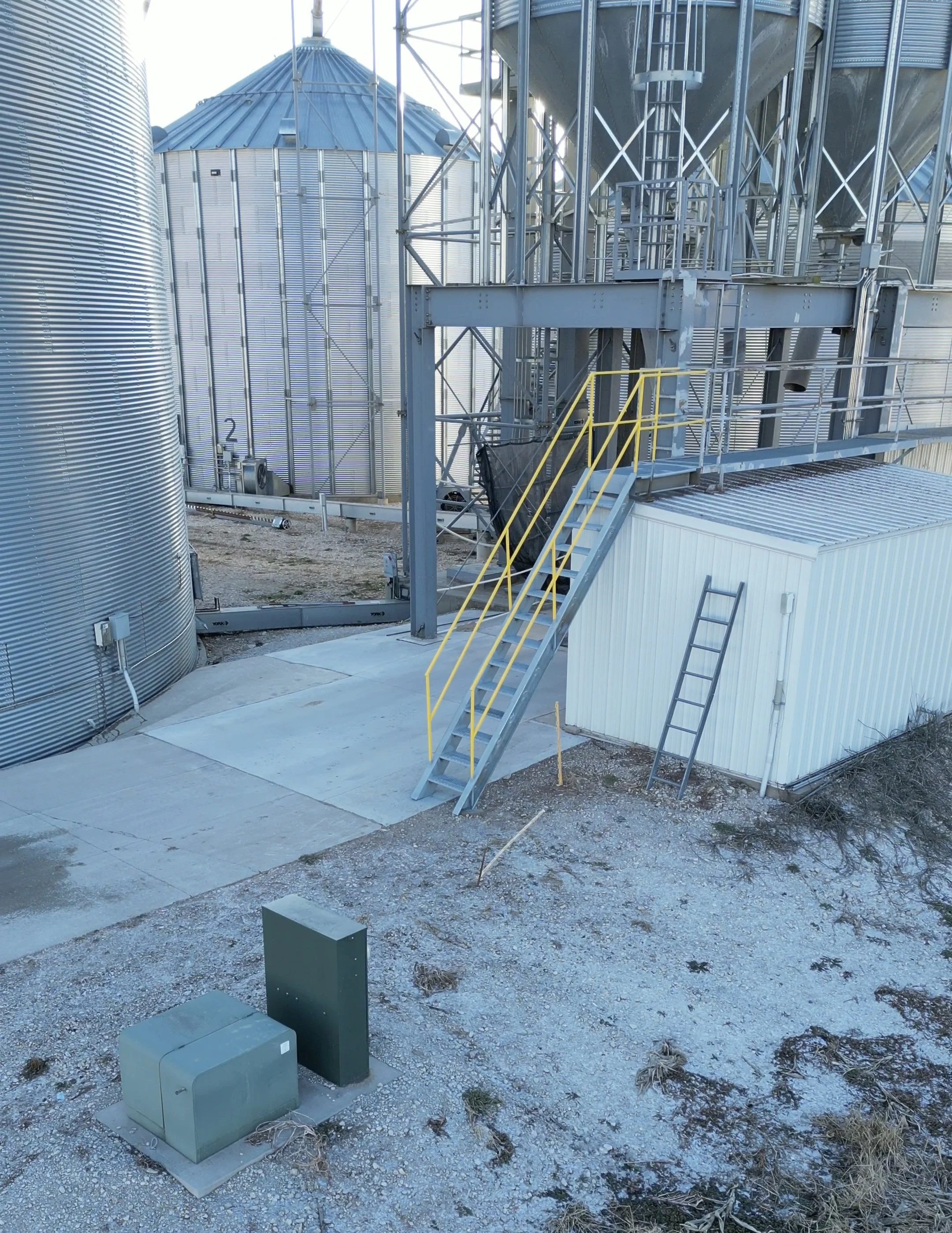 Industrial storage facility with metal silos and a metal staircase with yellow railings leading to an upper platform. A ladder leans against a white shed in the foreground. The ground is a mix of gravel and concrete with an electrical box and utility meters.