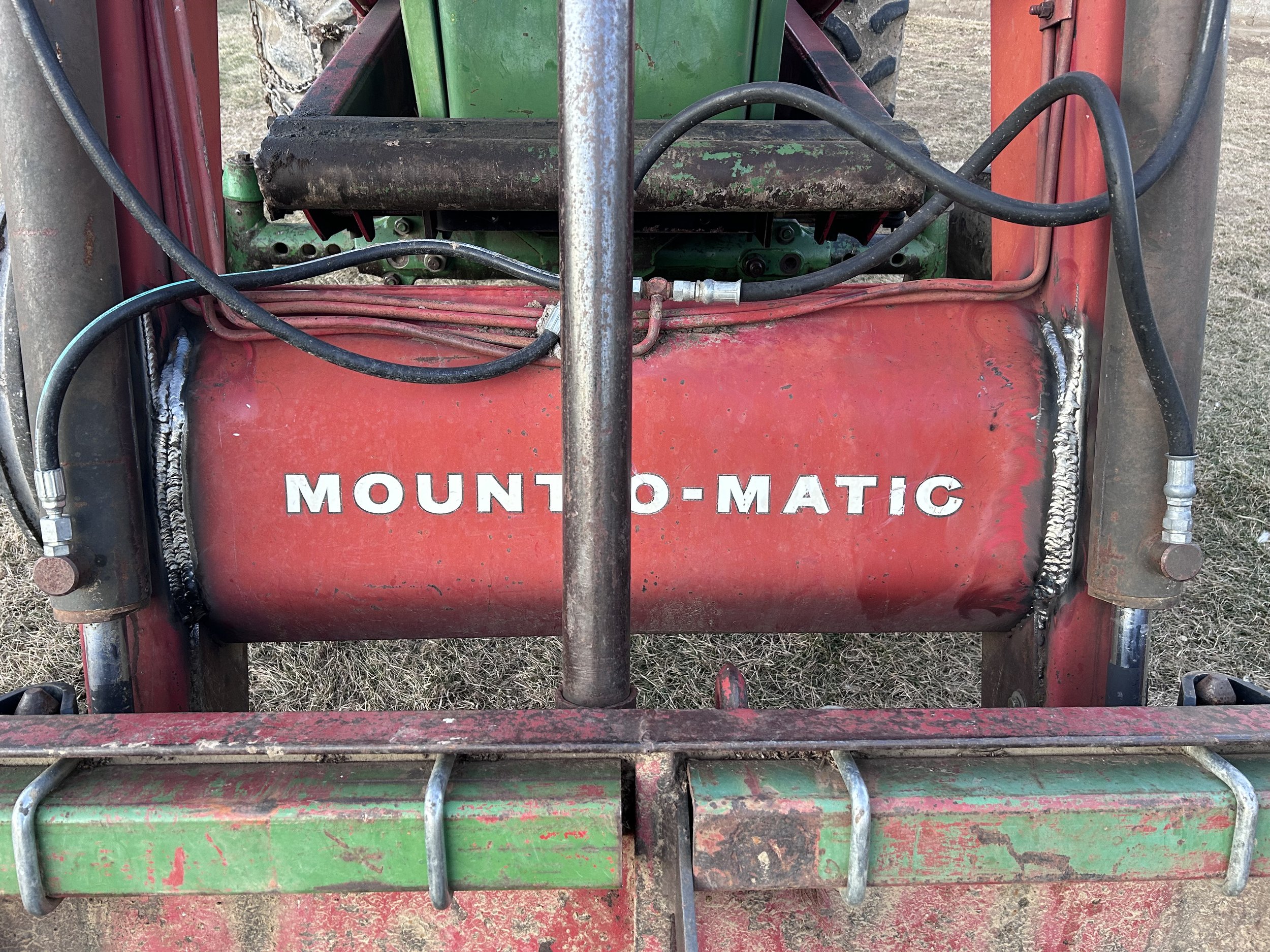 Close-up of a red Mounto-Matic fuel tank with welding and hoses attached, mounted on a green and red piece of machinery, showing signs of wear and dirt.