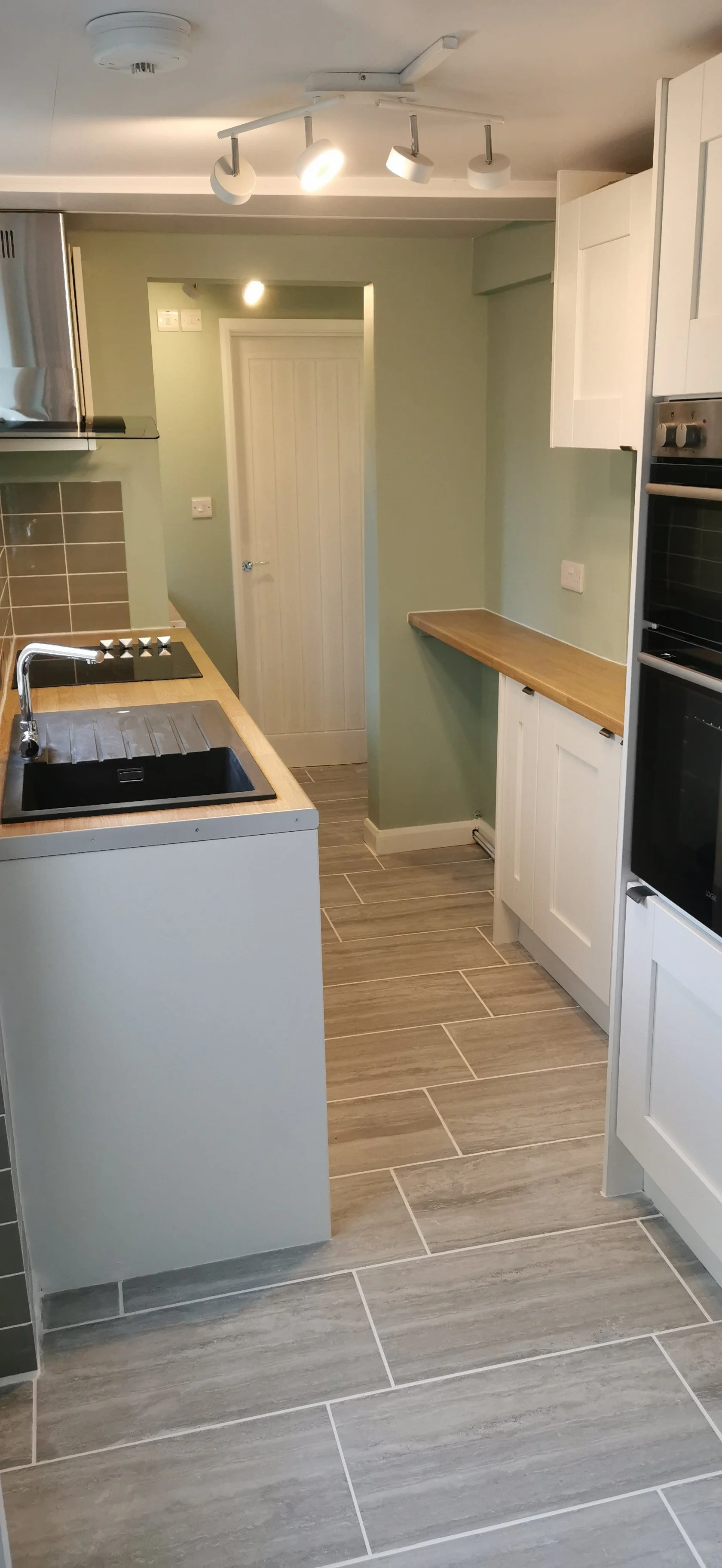 A modern kitchen with a light green wall, wooden countertops, white cabinets, a black sink, and grey floor tiles. There are overhead lights and a door at the back of the room.