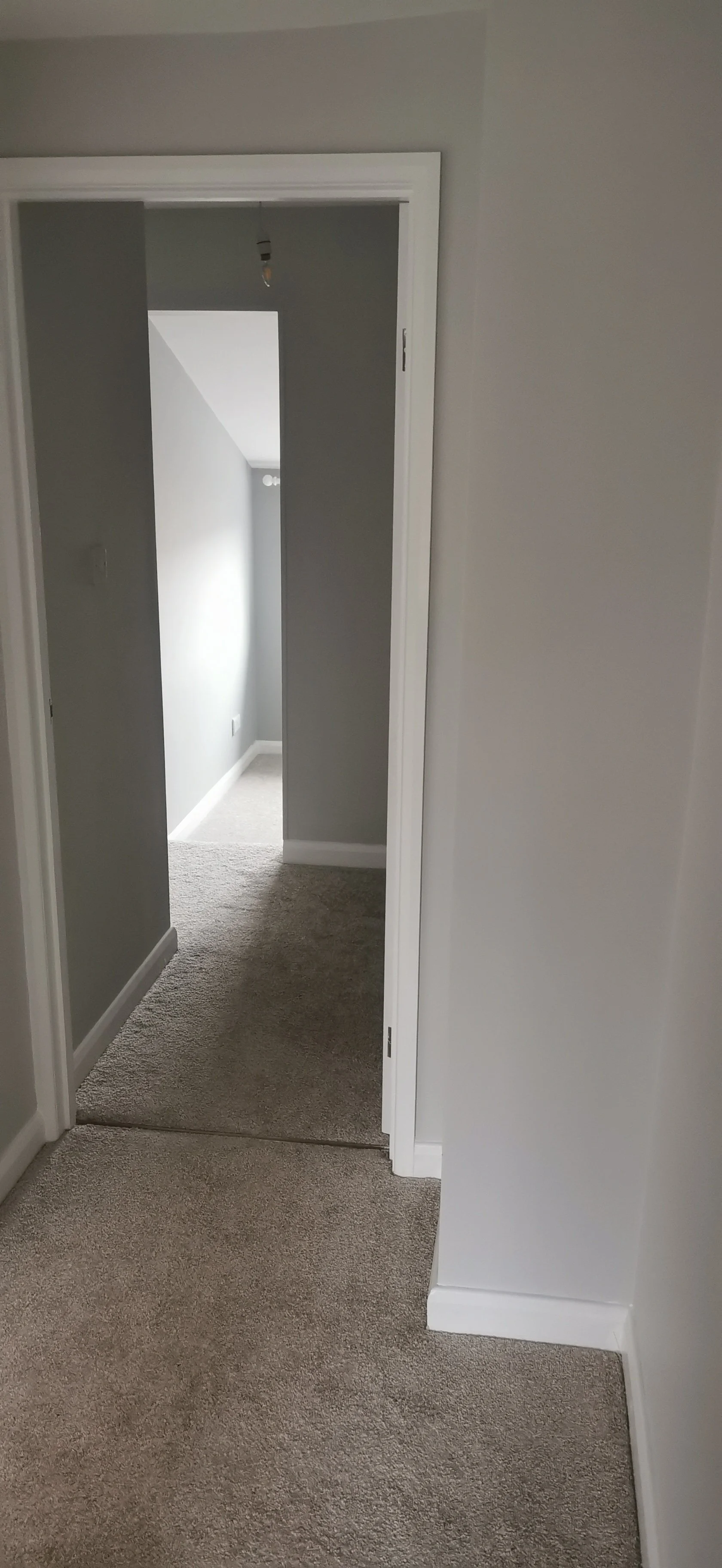 Empty room with doorway, gray walls, carpeted floor, and an exposed light bulb hanging from the ceiling.