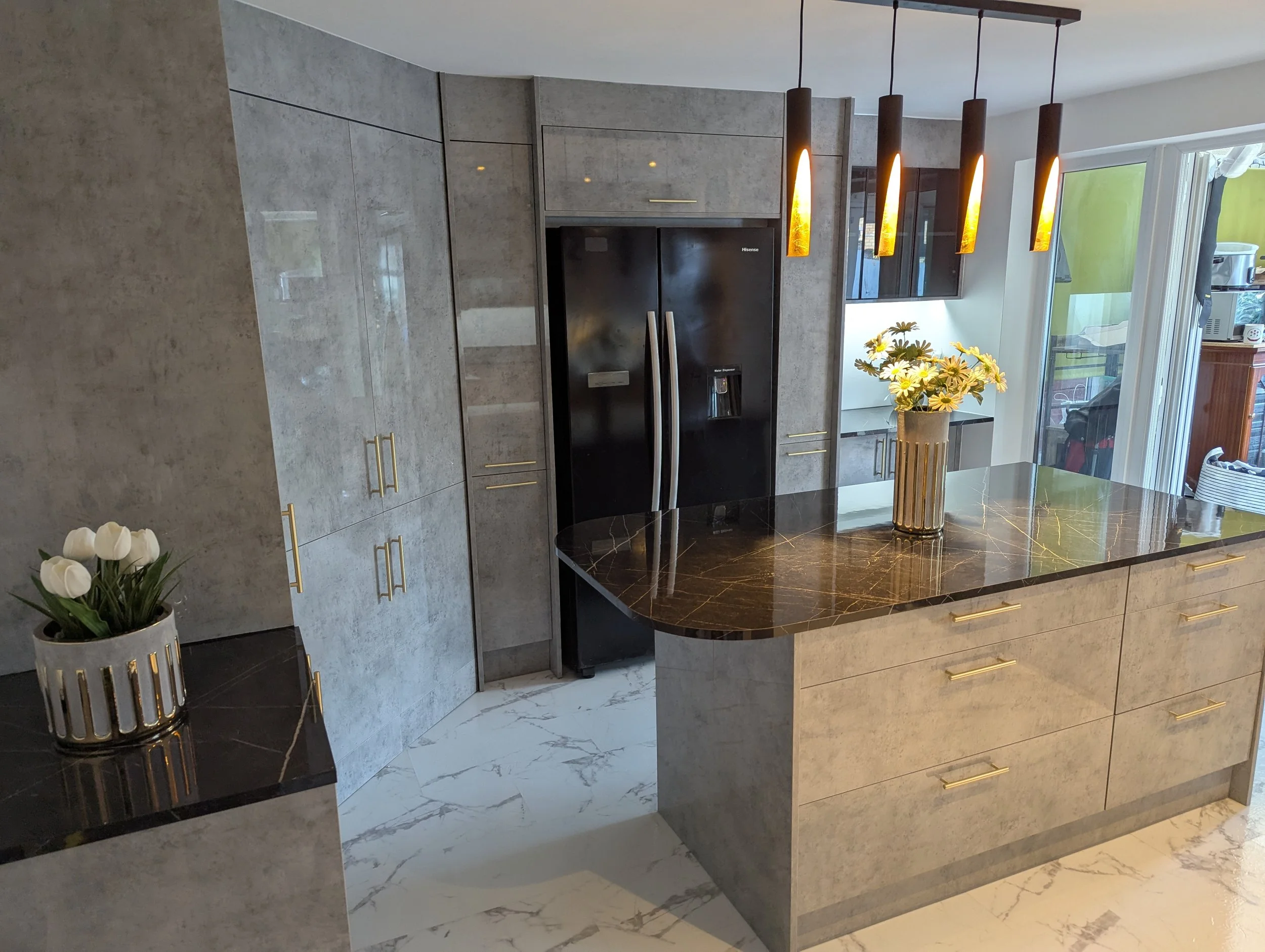 Modern kitchen with gray cabinetry, black refrigerator, marble island with gold handles, hanging pendant lights, and flower vases with white tulips and yellow daisies.