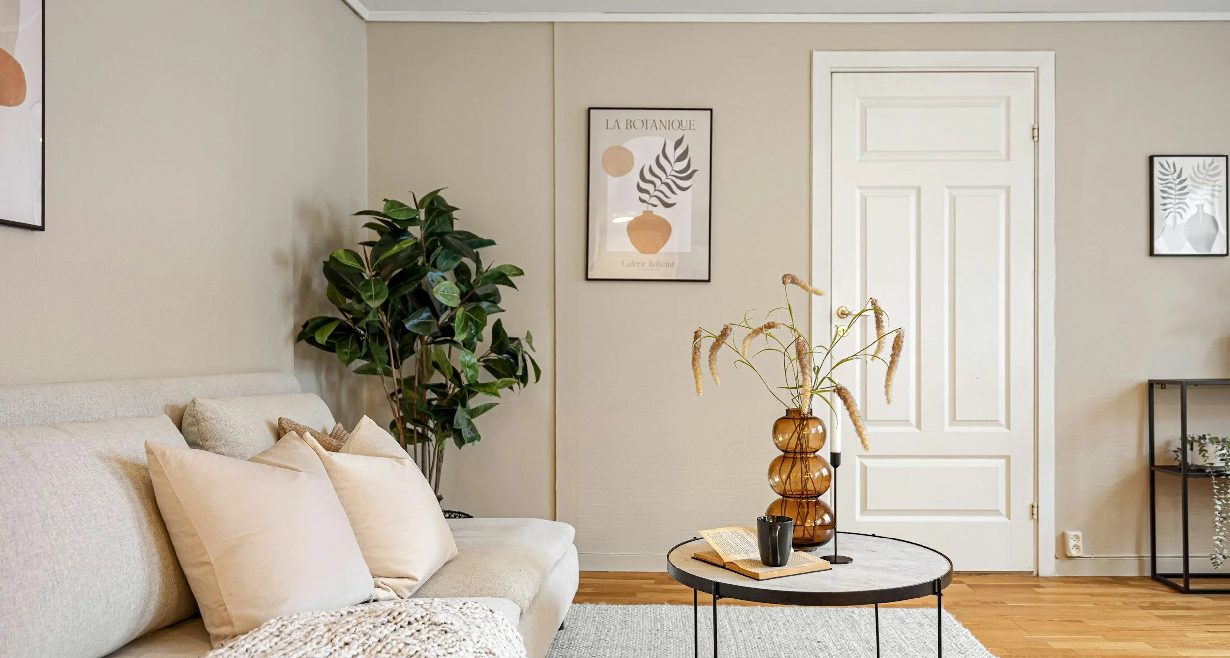 Living room with beige sofa, cream pillows, black round coffee table with book, black mug, brown vase with dried pampas grass, large green plant, framed art on beige walls, white door, and a black metal shelf with small plant.