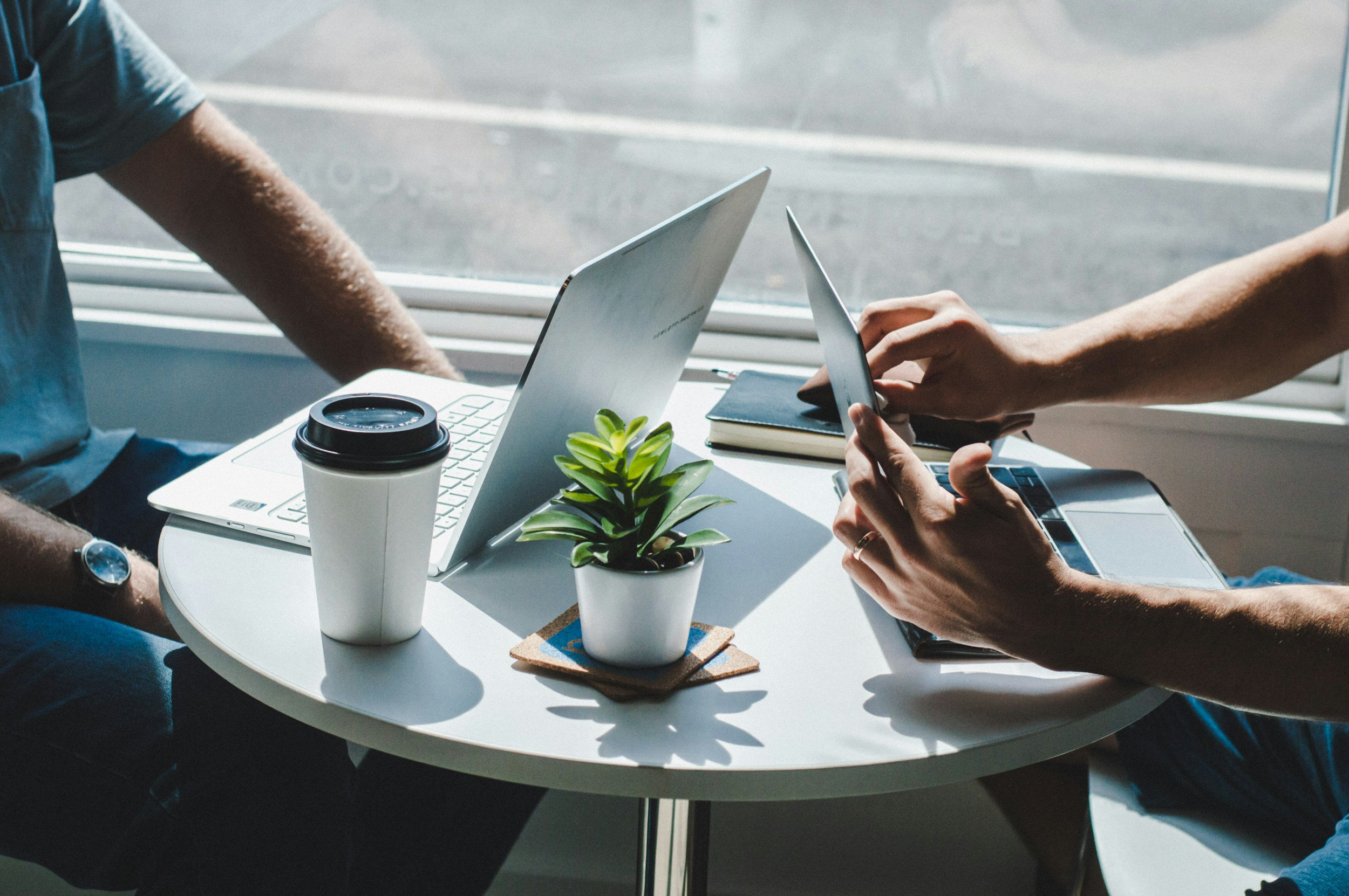 Two people working at a small round table in a coffee shop or office, with laptops, a coffee cup, a small potted plant, a notebook, and a tablet.