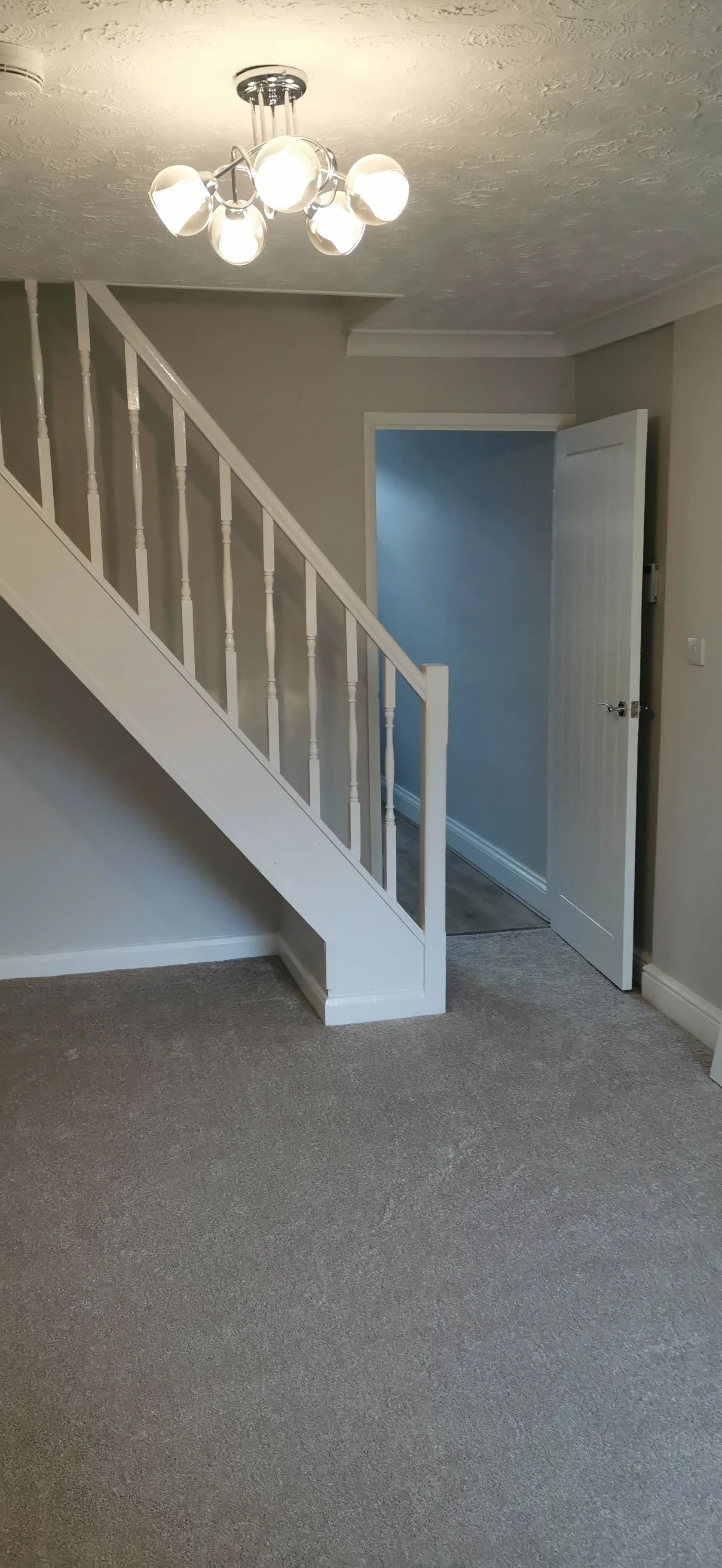 Interior room featuring a staircase with white railing, a modern ceiling light fixture with multiple bulbs, gray walls, beige carpet, and an open door leading to another room.