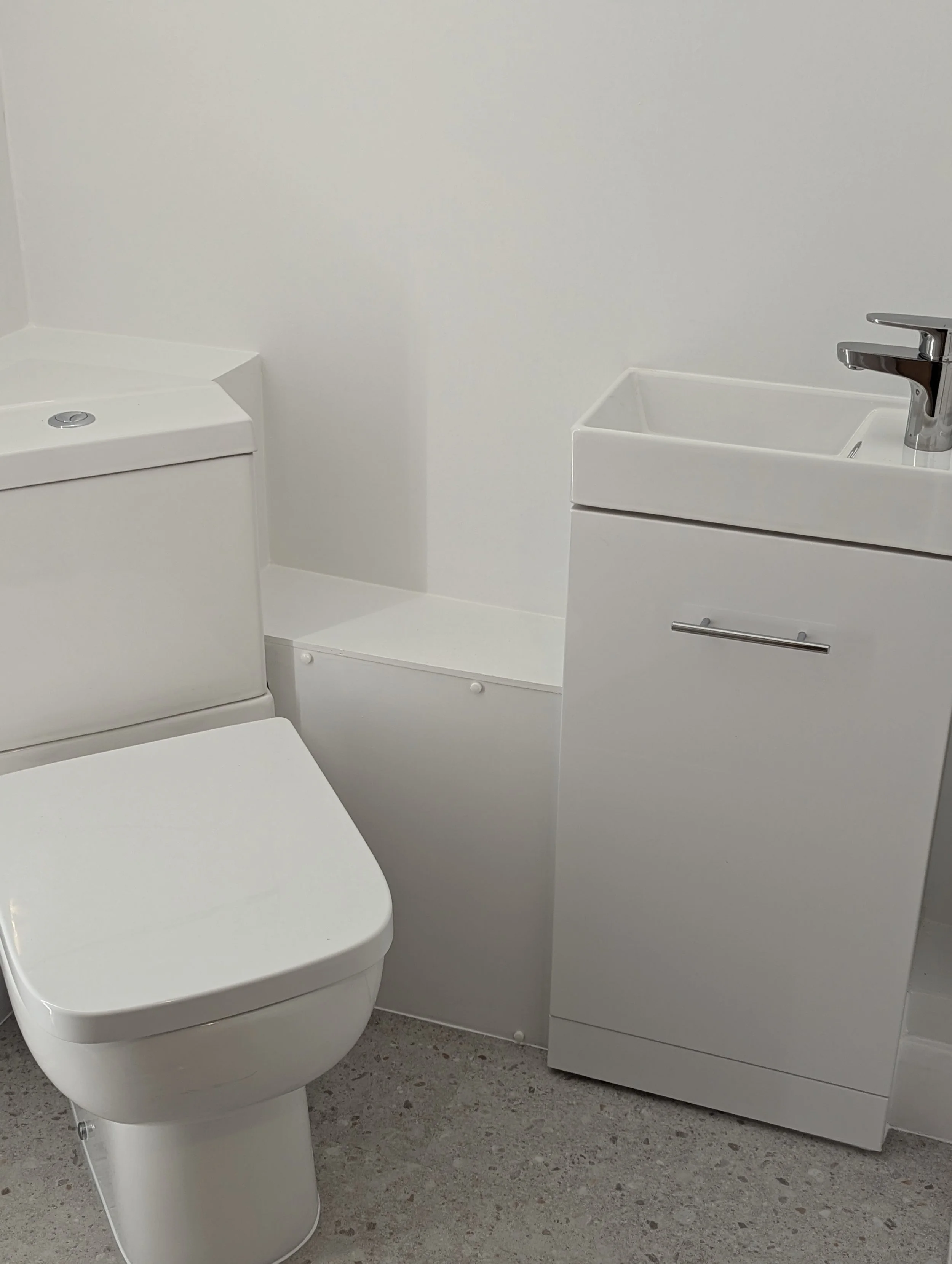 A small, modern bathroom with a white toilet and a white sink cabinet. The sink has a chrome faucet, and the bathroom has white walls and a speckled gray floor.