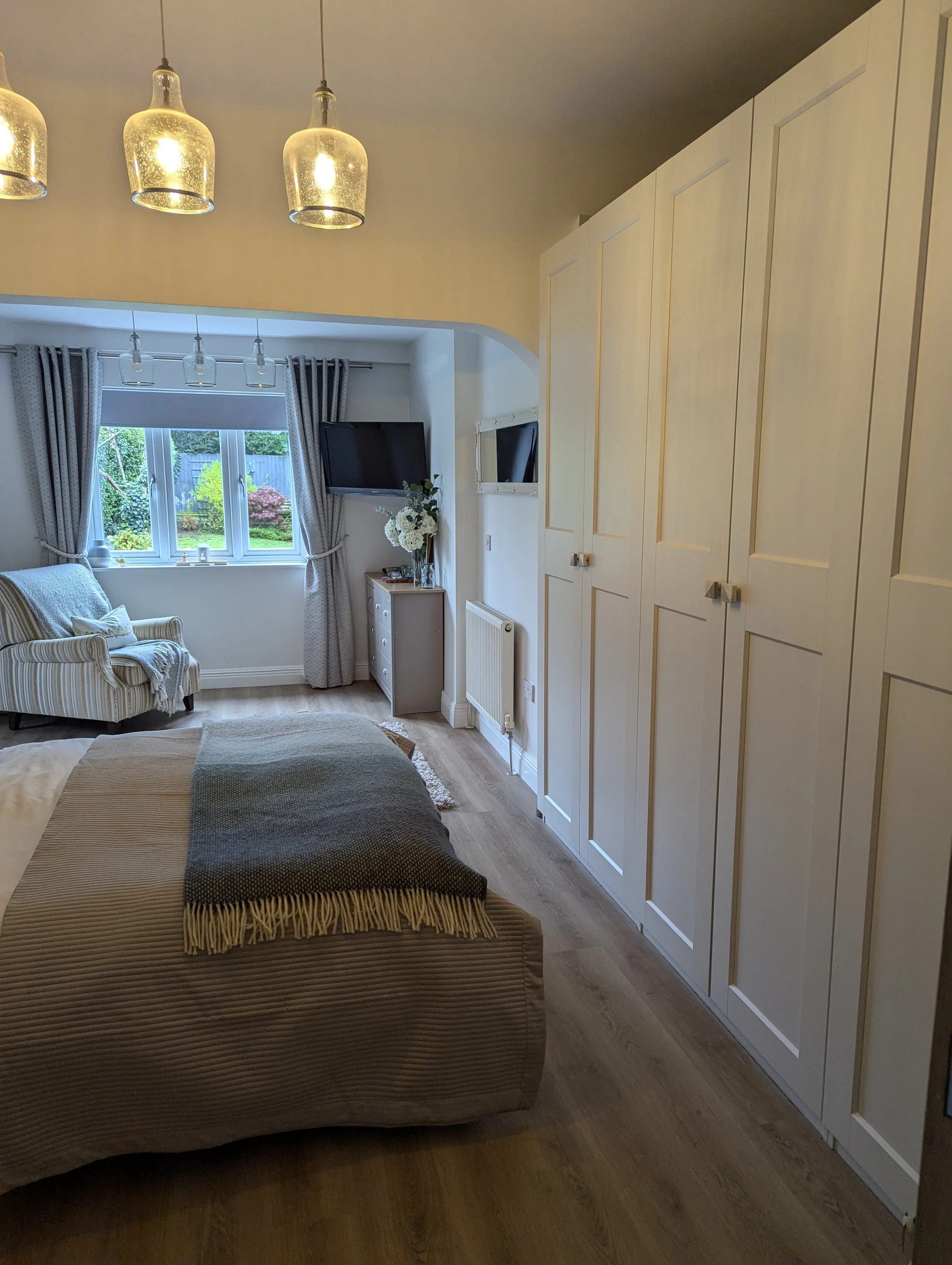 A cozy bedroom with a bed covered in a striped beige duvet, a cream-colored armchair with blue and beige stripes near a window with gray curtains, and a large white wardrobe on the right side. There is a flat-screen TV mounted on the wall, a small ch