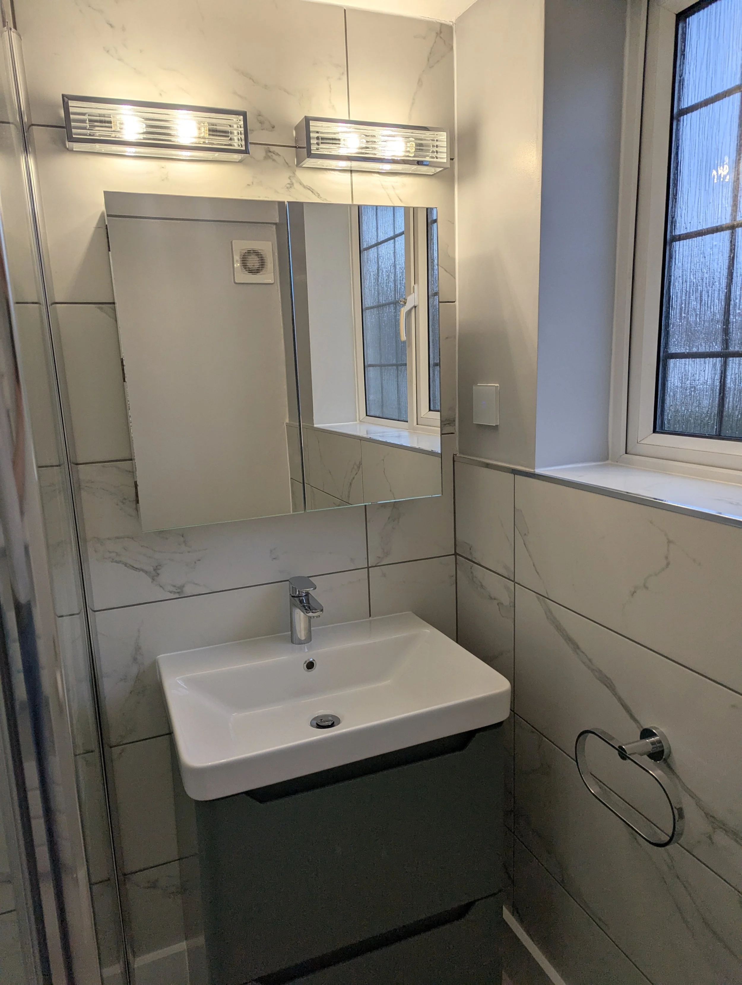A small bathroom with marble tiles, a rectangular mirror, a wall-mounted sink with a chrome faucet, a window with frosty glass, and a wall-mounted light fixture above the mirror.