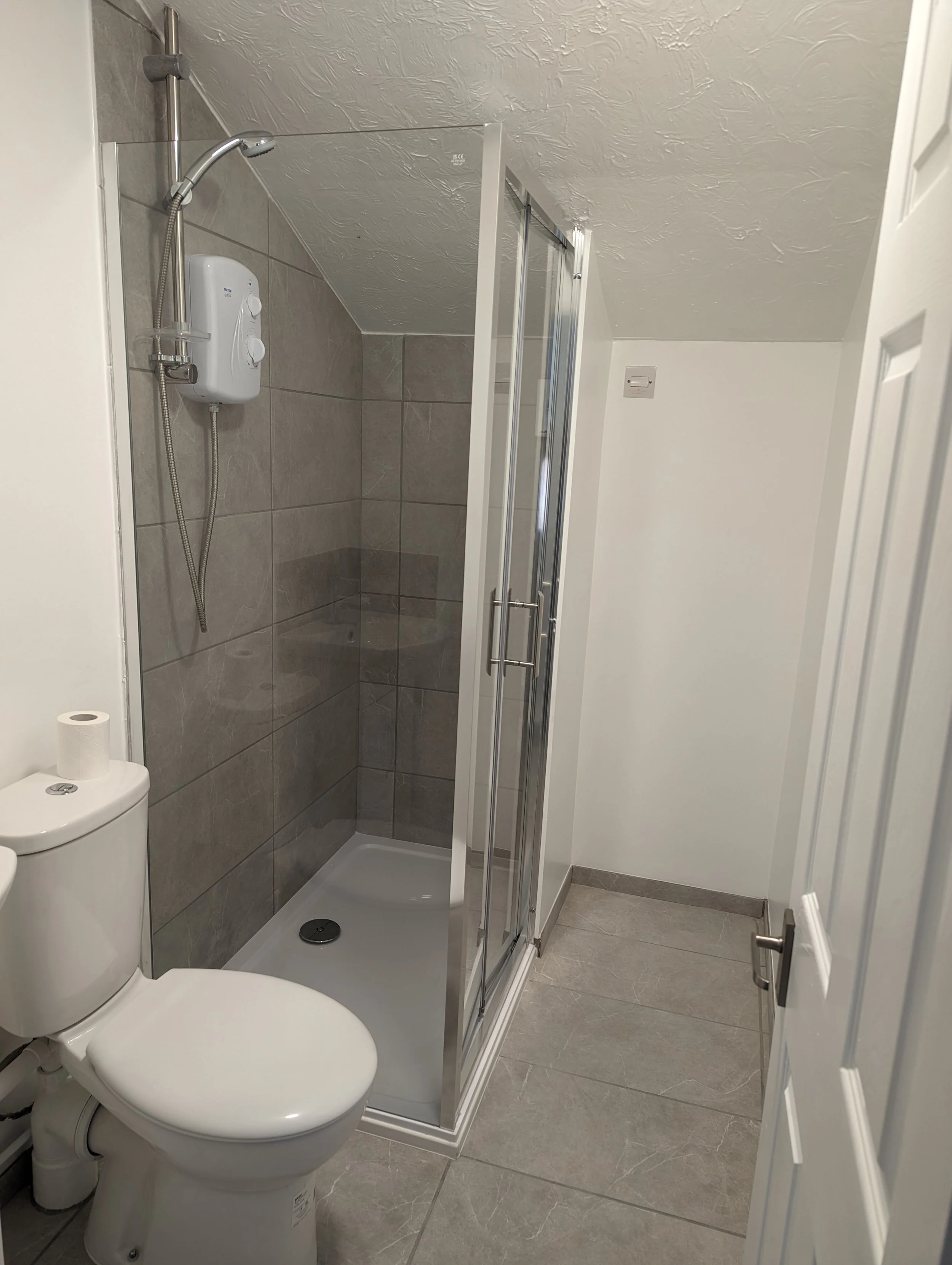 Small bathroom with toilet, shower stall with glass door, and gray tiled walls and floor.