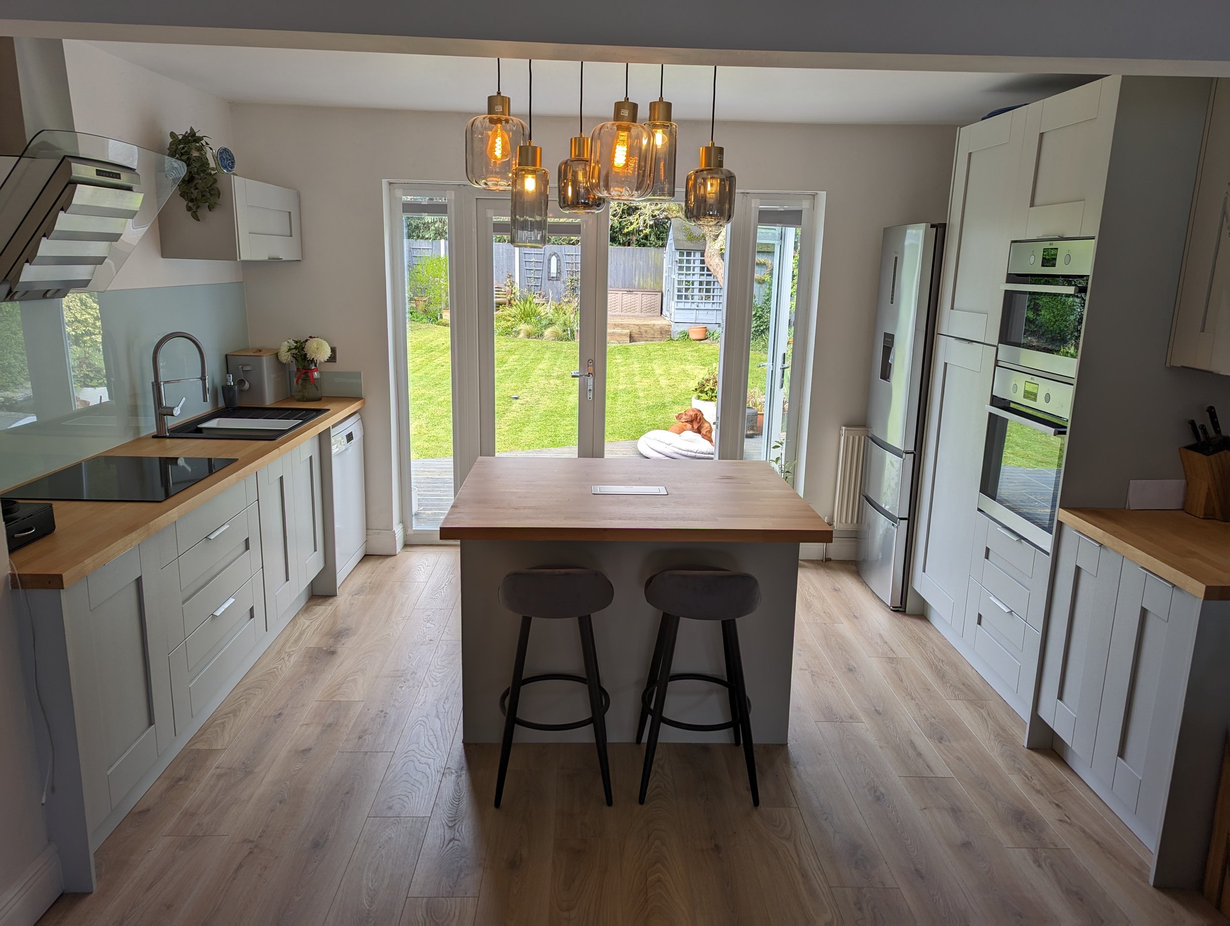 Modern kitchen with white cabinetry, wooden countertops, and a central island with two stools. Large glass doors lead to a backyard with green grass and a dog resting on a cushion.
