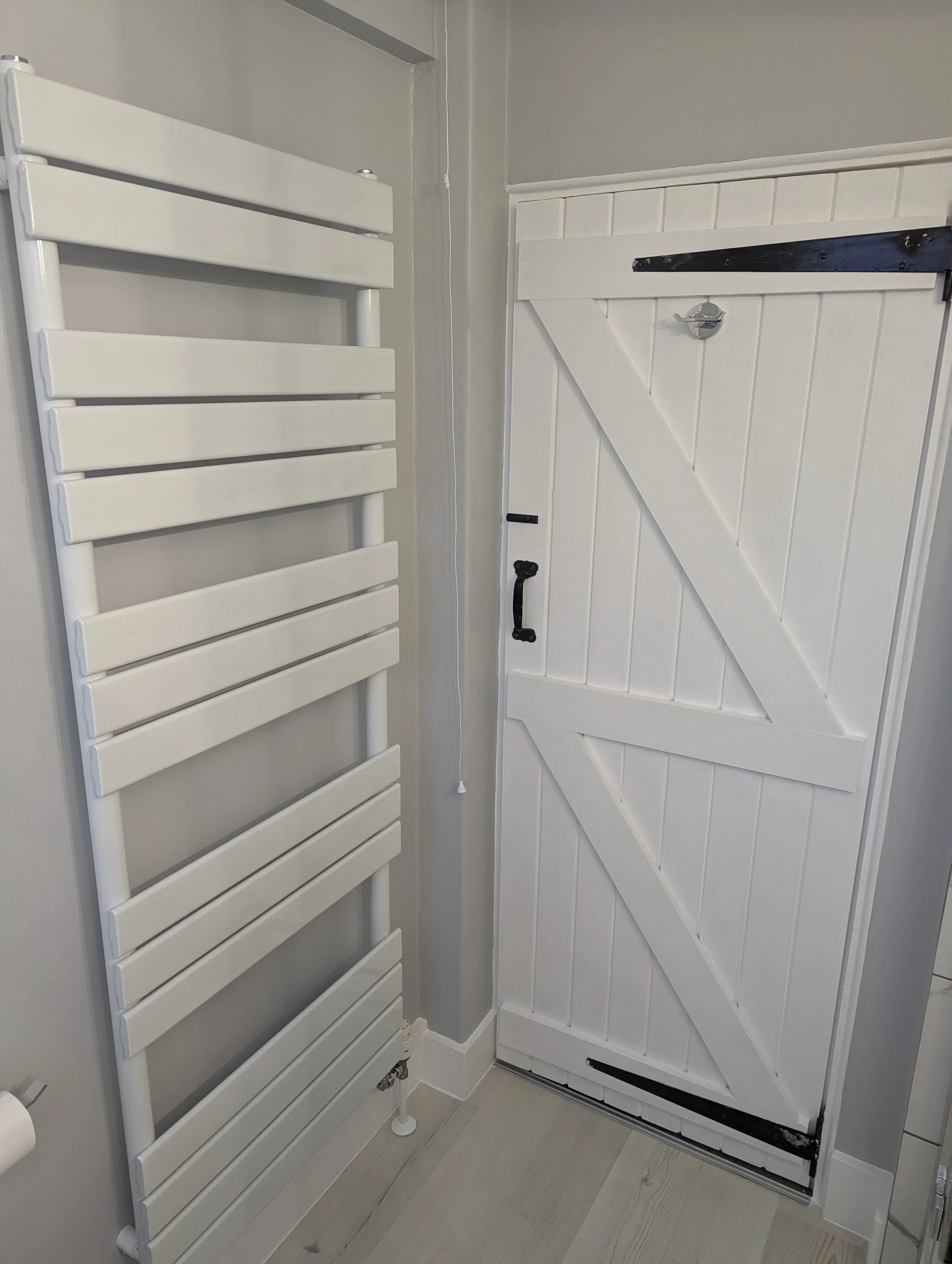 A white heated towel rail next to a white wooden barn door with black hardware, situated in a modern bathroom with gray walls and light-colored flooring.