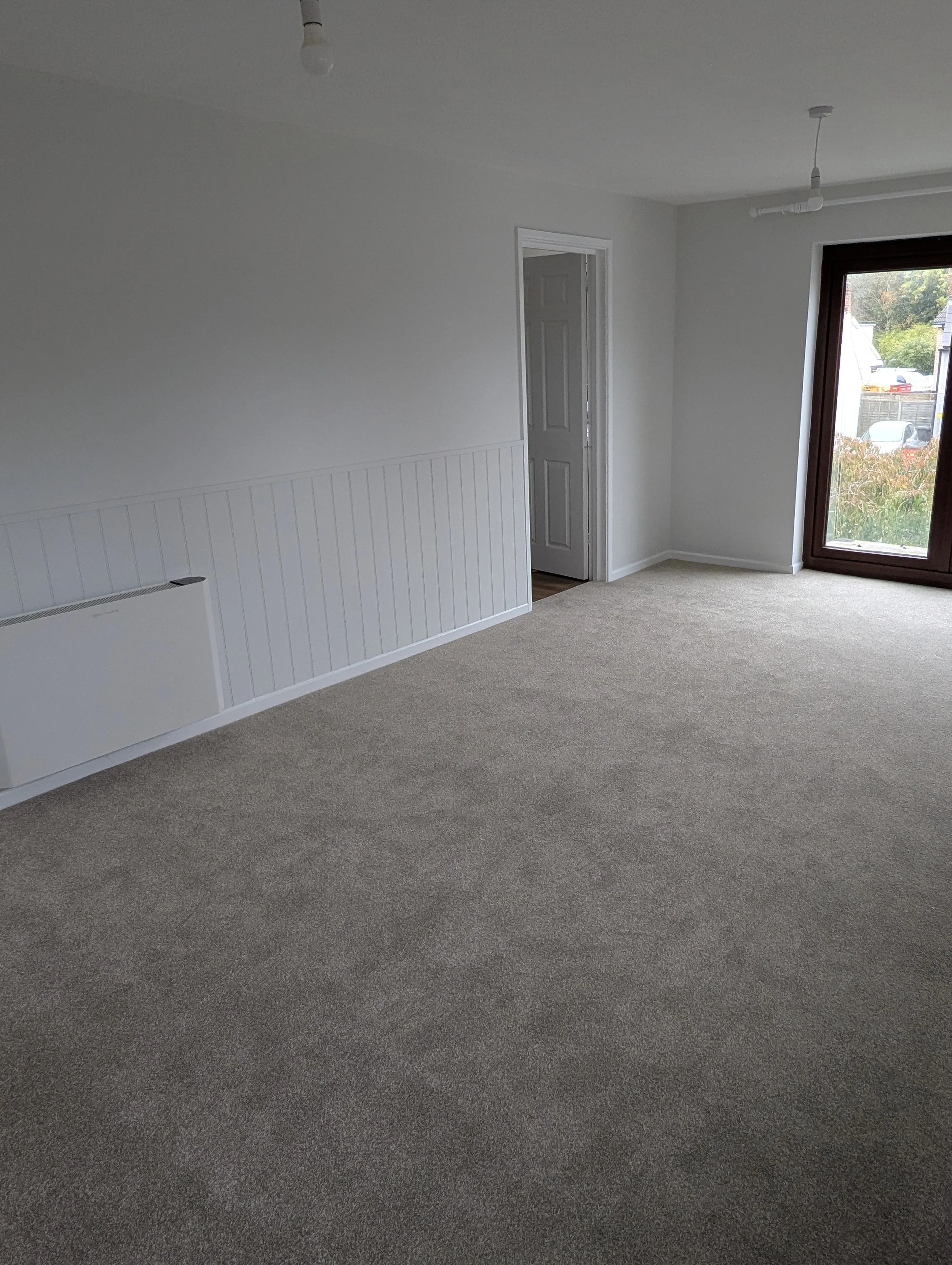 Empty living room with gray carpet, white walls, a door, a large window, and a wall-mounted heater.