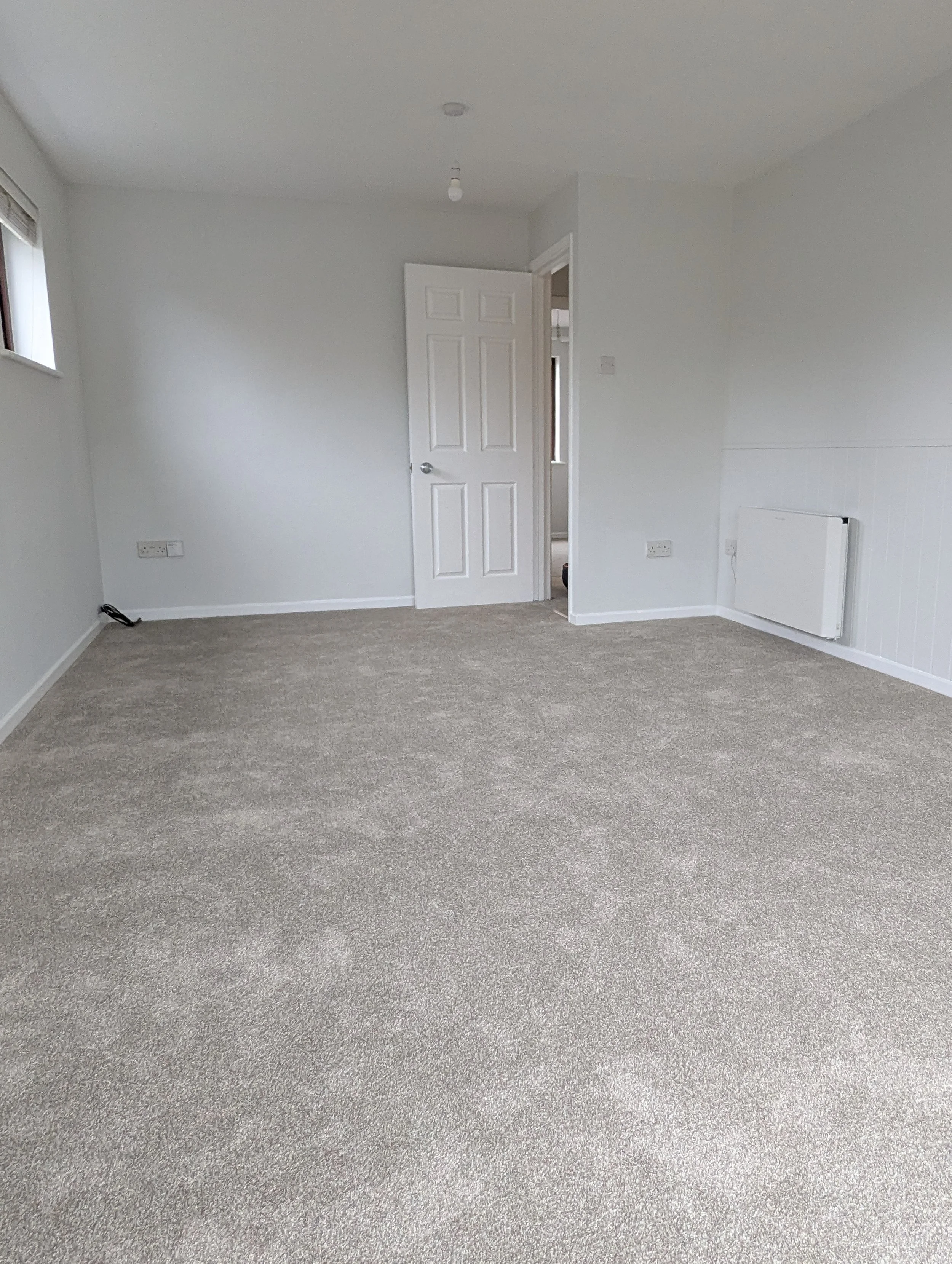 Empty beige-carpeted room with white walls, a white door slightly open, a small window, and a white radiator.